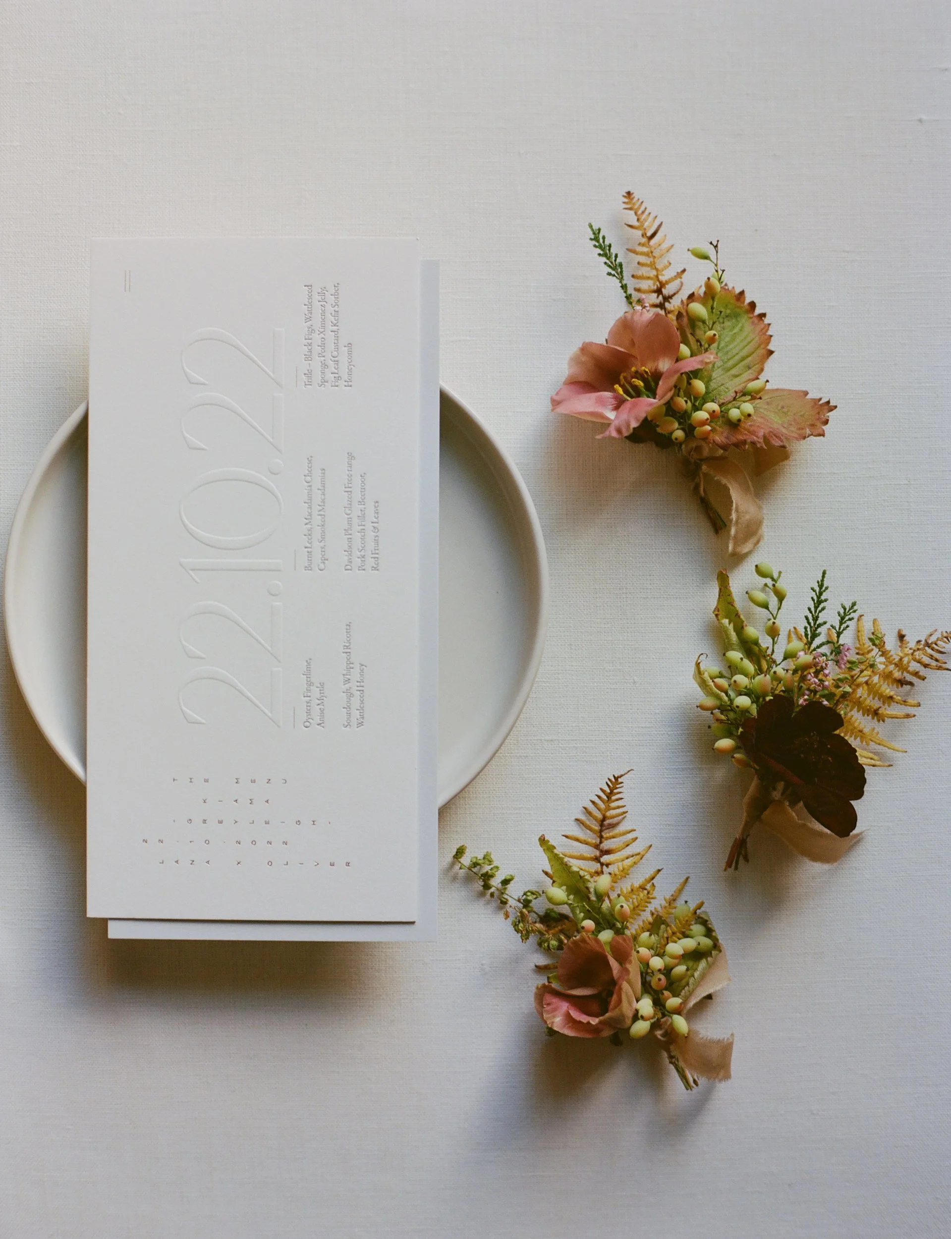 Wedding invitation on a white plate with three boutonnières of flowers and greenery on a white table.