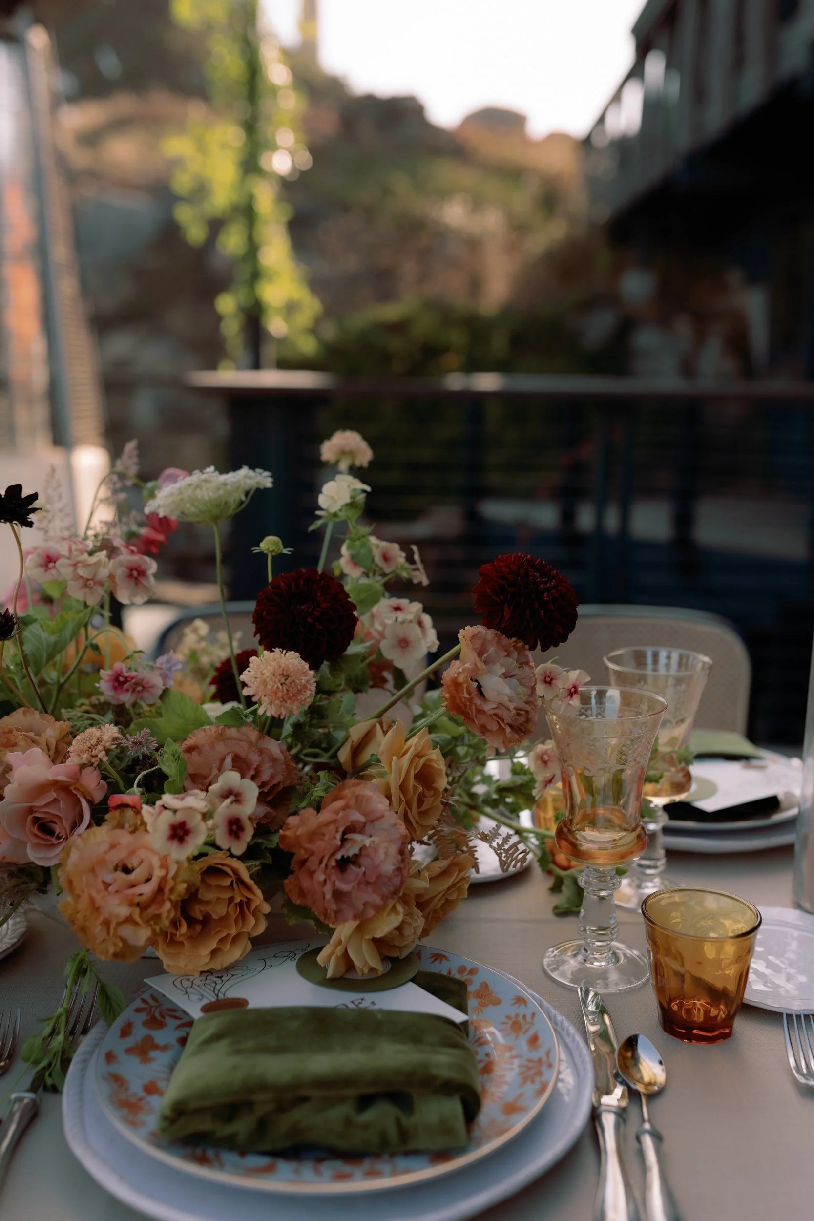 A table set outdoors with a floral centerpiece, colorful glassware, and a plate with a green cloth napkin.