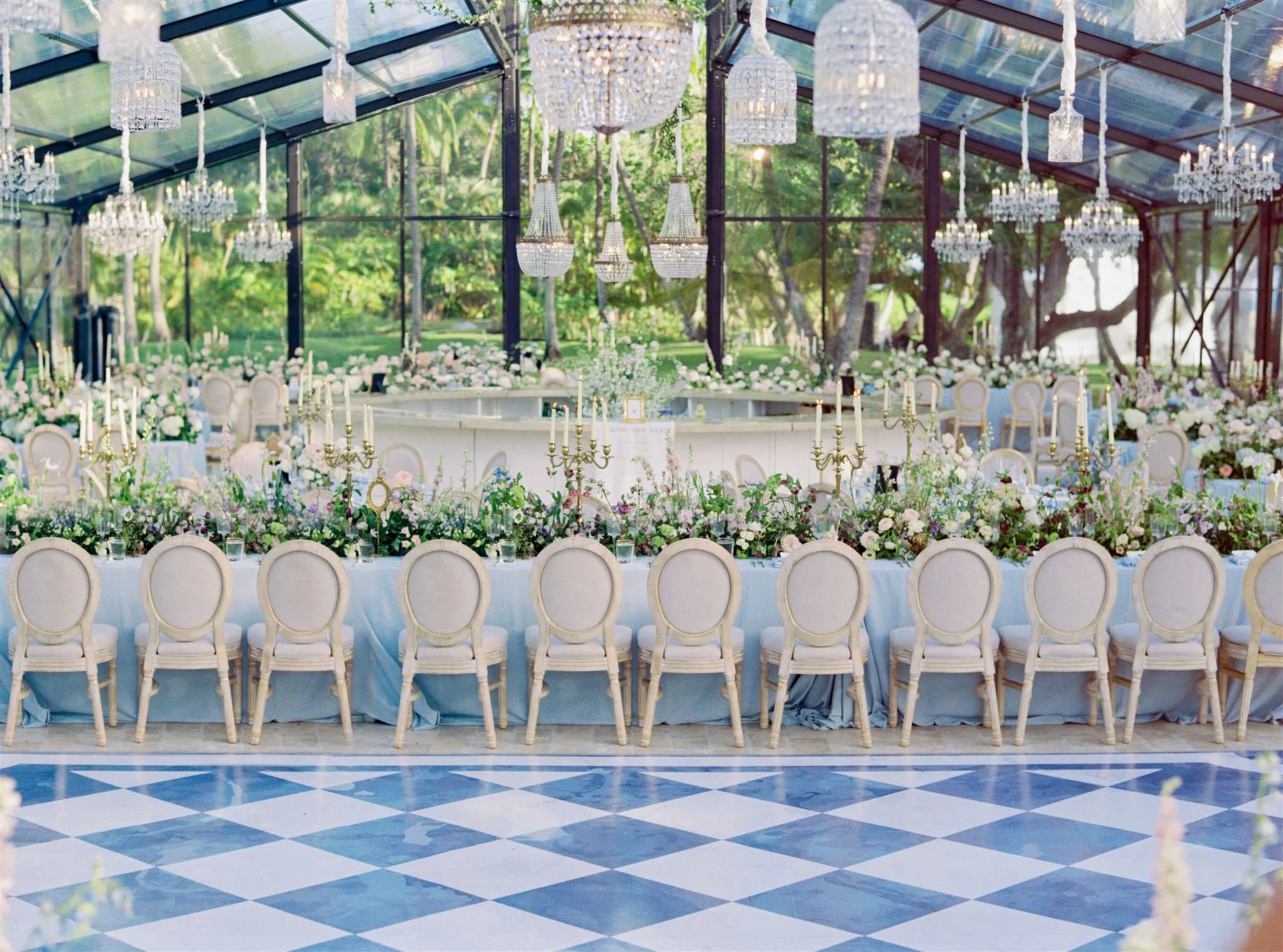 Elegant banquet setup in a glass-roofed conservatory with long tables, floral arrangements, gold candelabras, chandeliers, and white chairs, greenery visible outside.