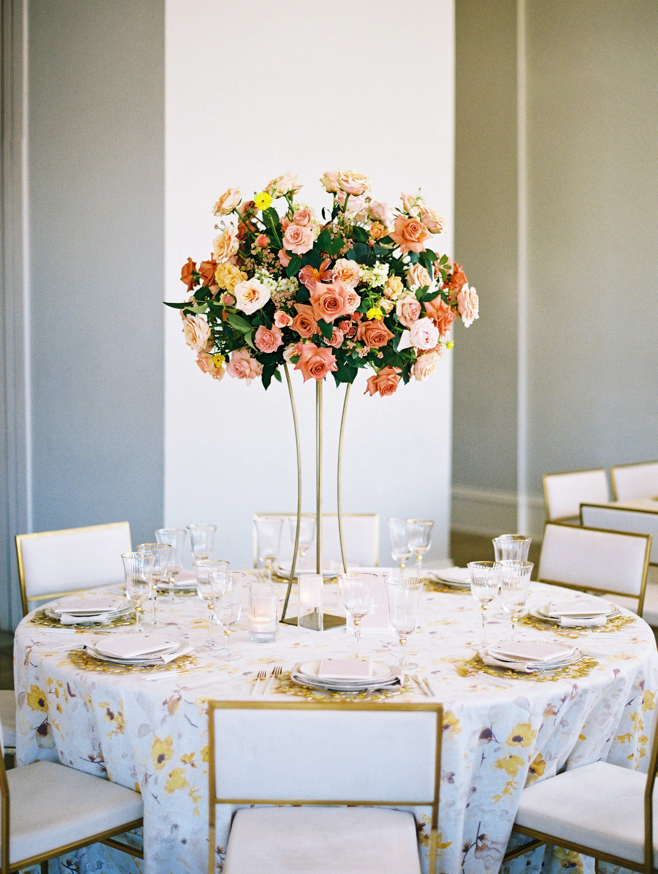 round table and chairs with a gold stand in the center with a round floral arrangement with peach, pink, and yellow flowers on top