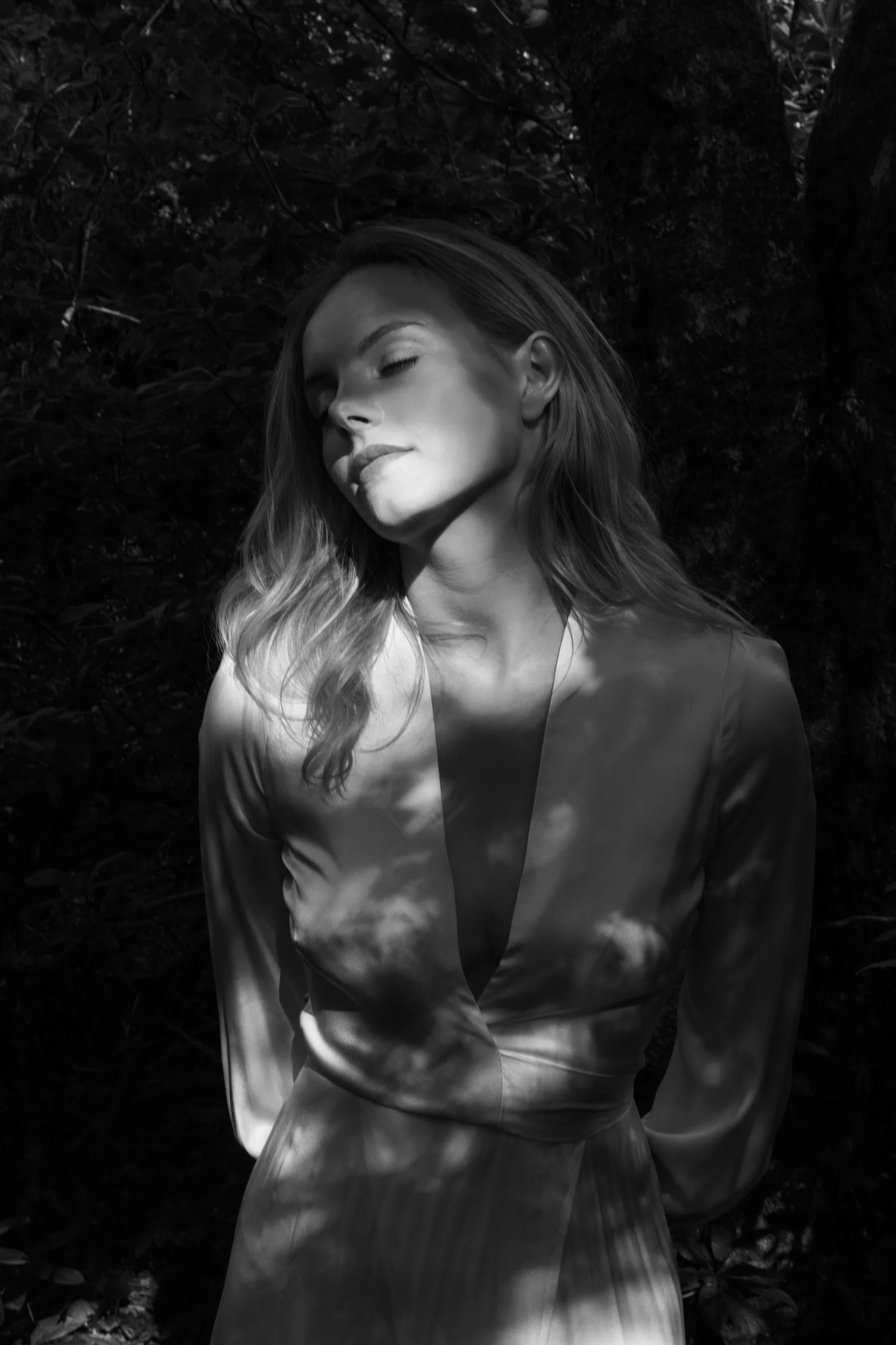 Black and white photo of a woman with long hair, closed eyes, and a serene expression, standing outdoors with shadow patterns on her face and dress.