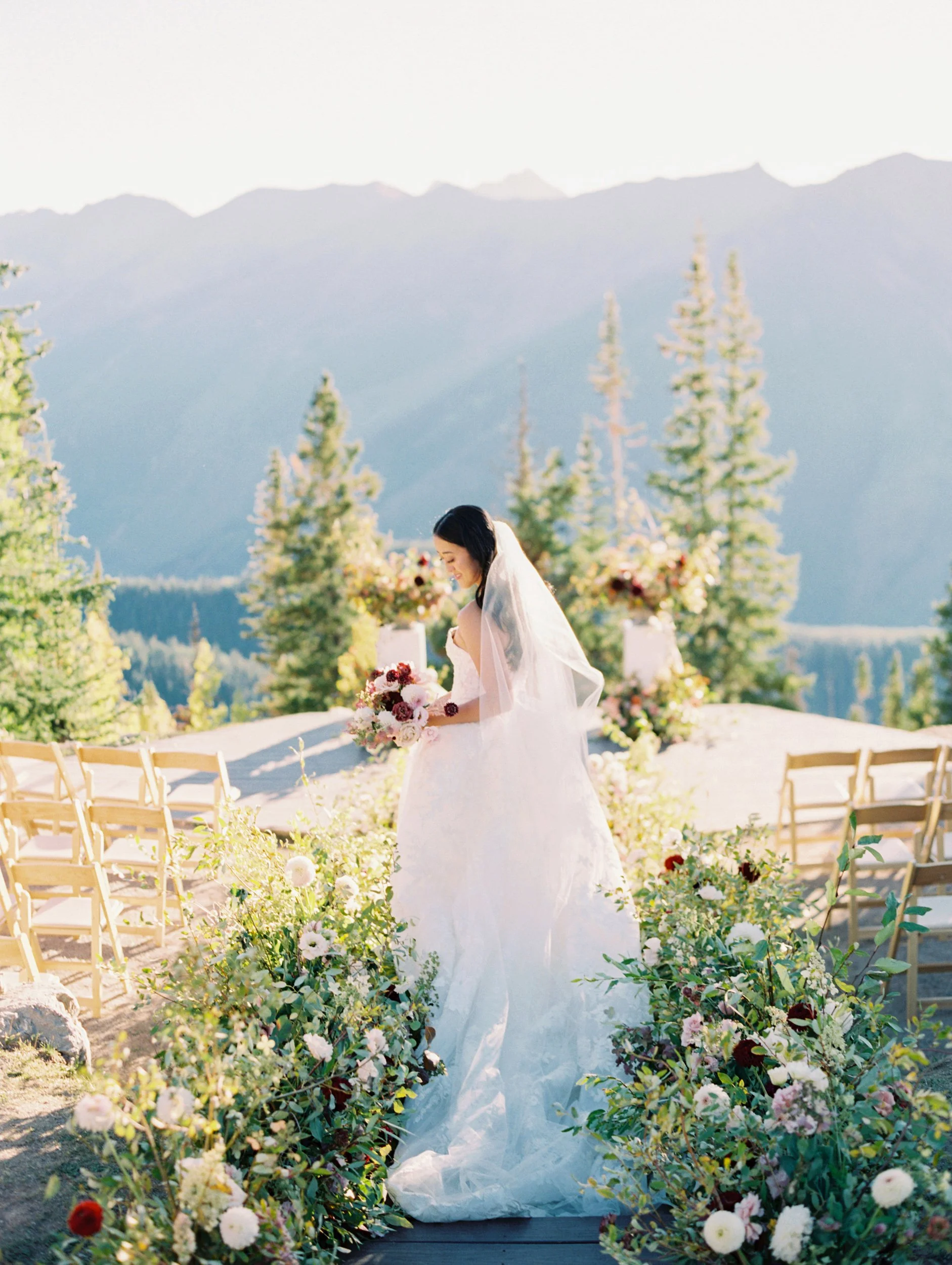 a bride looking down at her bouquet on top of a mountain with flowers surrounding her