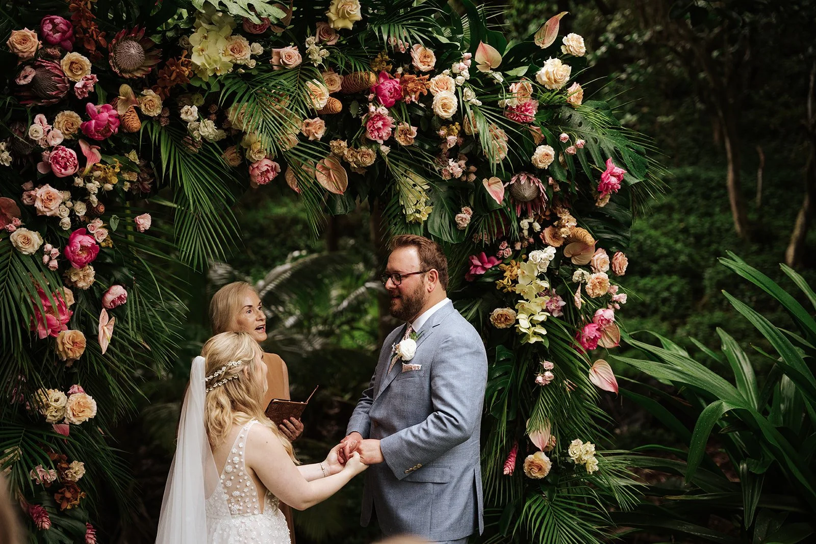 lady in white dress and main in grey suit holding hands under a floral and greenery arch