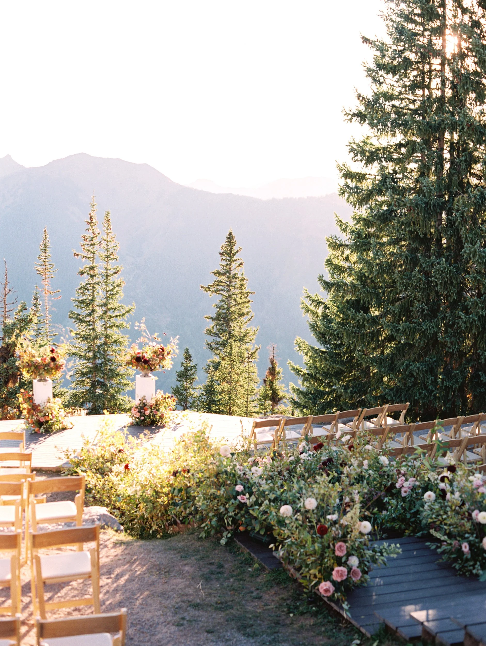 Outdoor wedding setup with rows of wooden chairs, floral arrangements, and mountain landscape in the background during sunset.