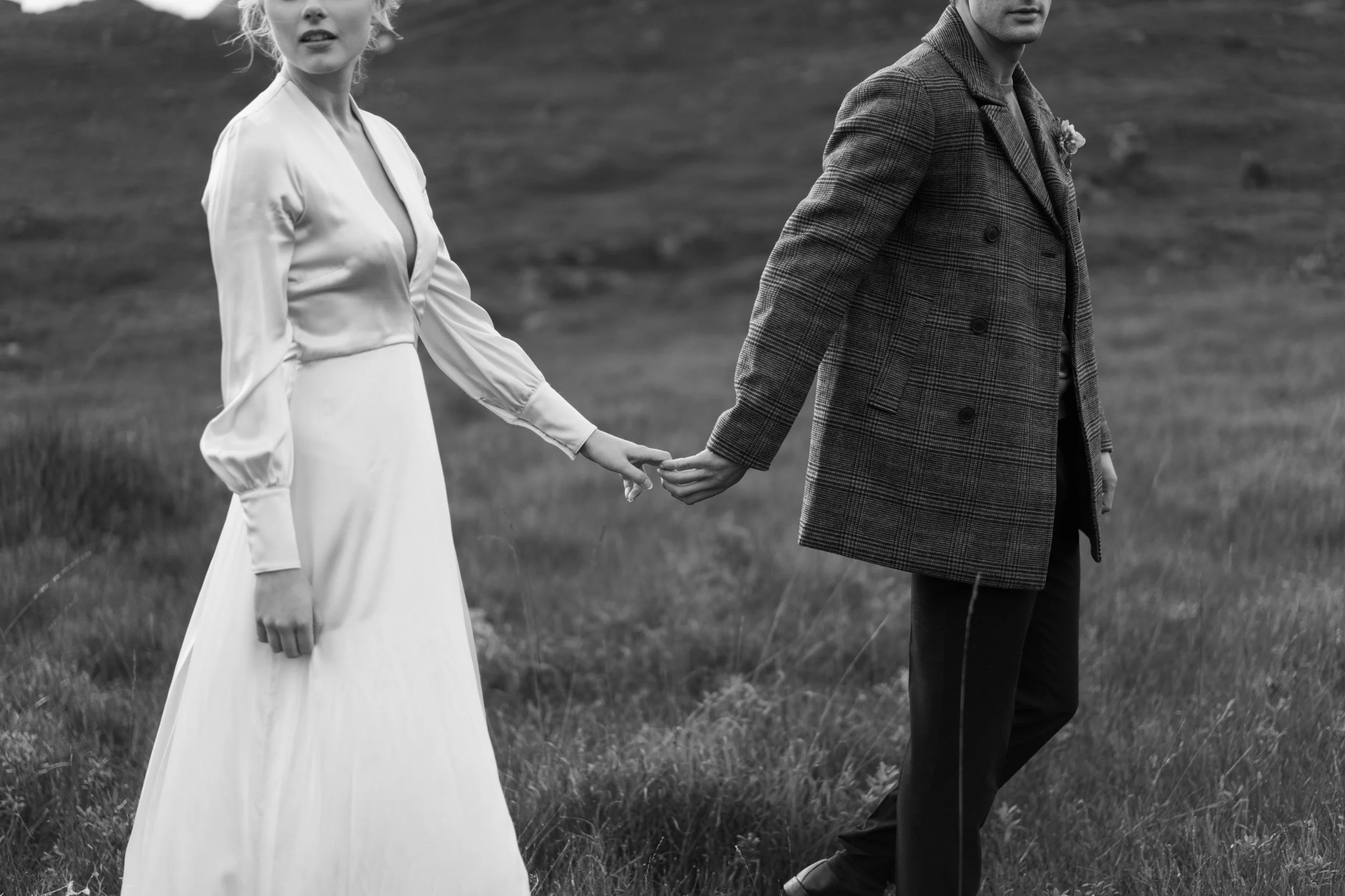 A black and white photo of a couple holding hands in a grassy field, with the woman wearing a long dress and the man wearing a plaid coat.