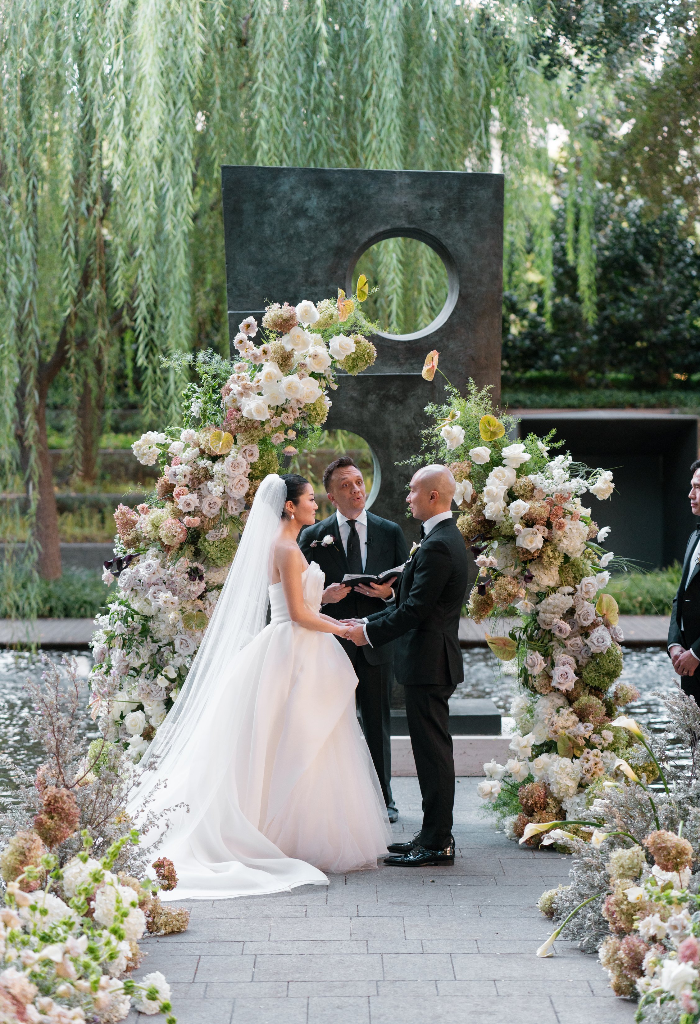 Bride and groom saying vows in front of lush floral arch at Nasher Sculpture Center