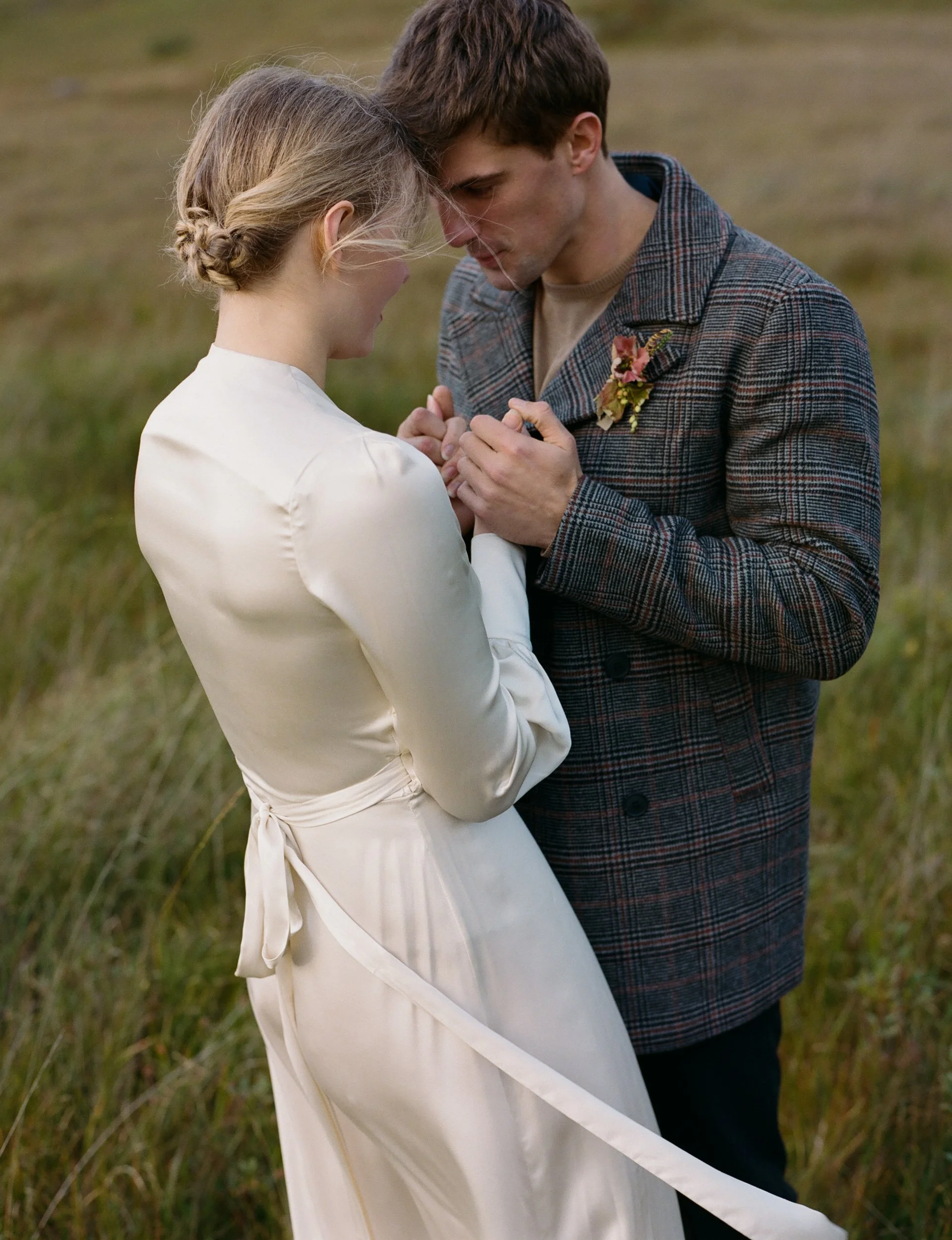 A couple dressed in vintage wedding attire holding hands and touching foreheads in a grassy field.