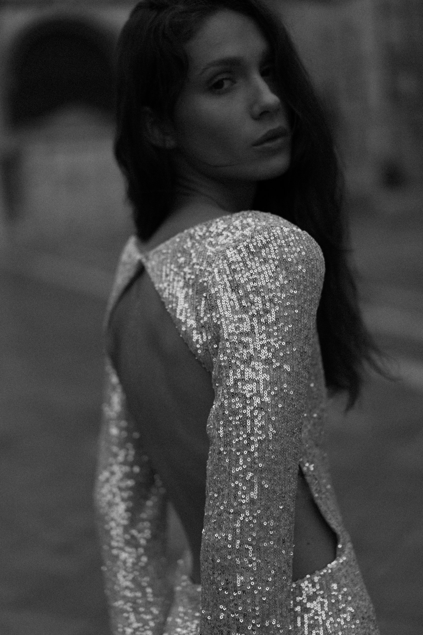 A woman with long dark hair turned to the side, wearing a shiny, sequined dress with open back details.
