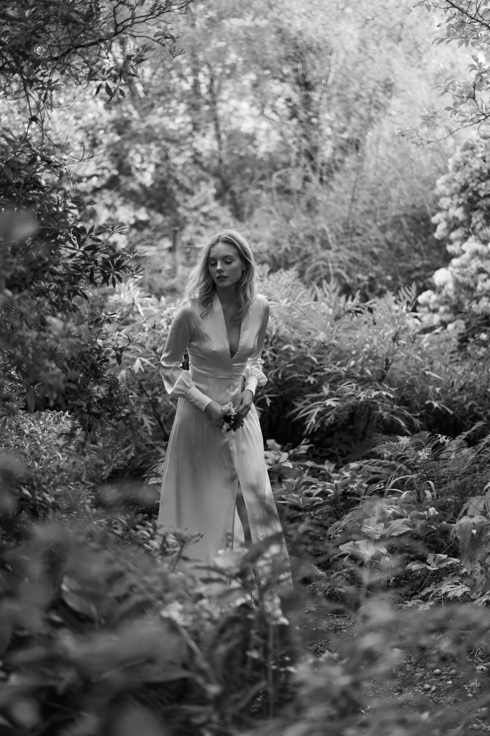 A woman in a long, elegant dress standing among lush, leafy vegetation in a natural outdoor setting, holding a small bouquet of flowers.