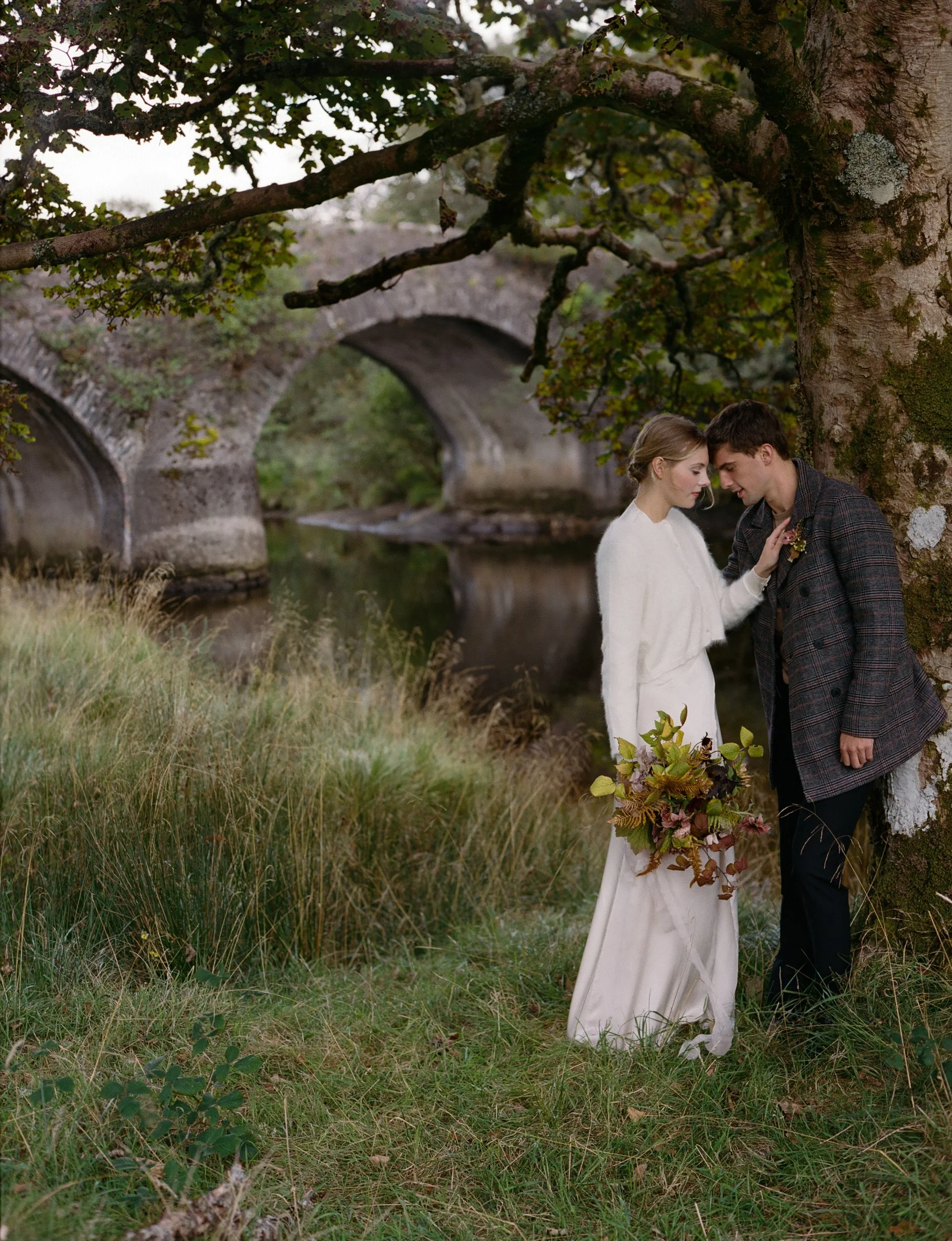 A couple on their wedding day standing under a large tree near a river with an old stone bridge in the background.