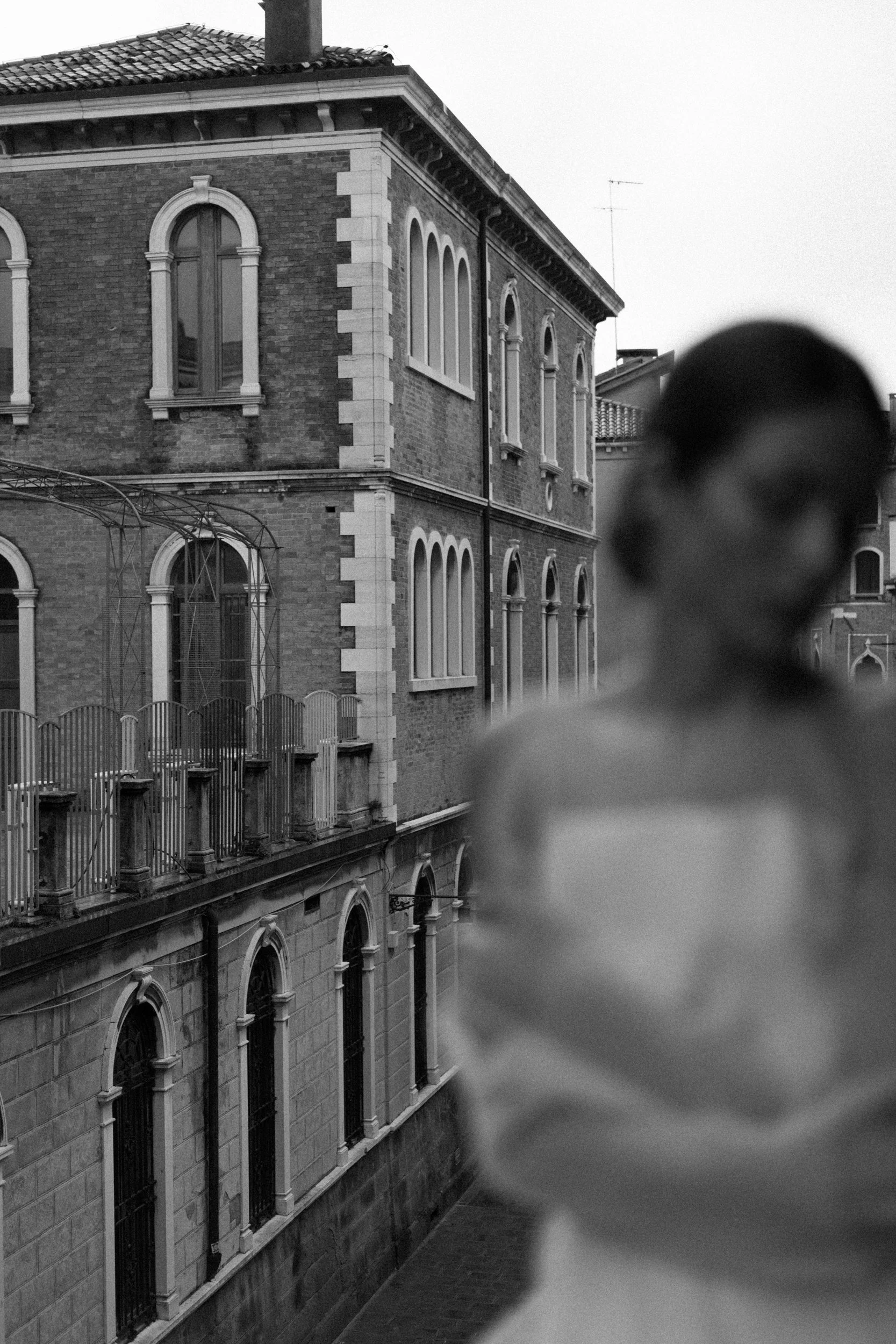 Black and white photo of a woman looking away, standing in front of an old brick building with arched windows and a balcony.