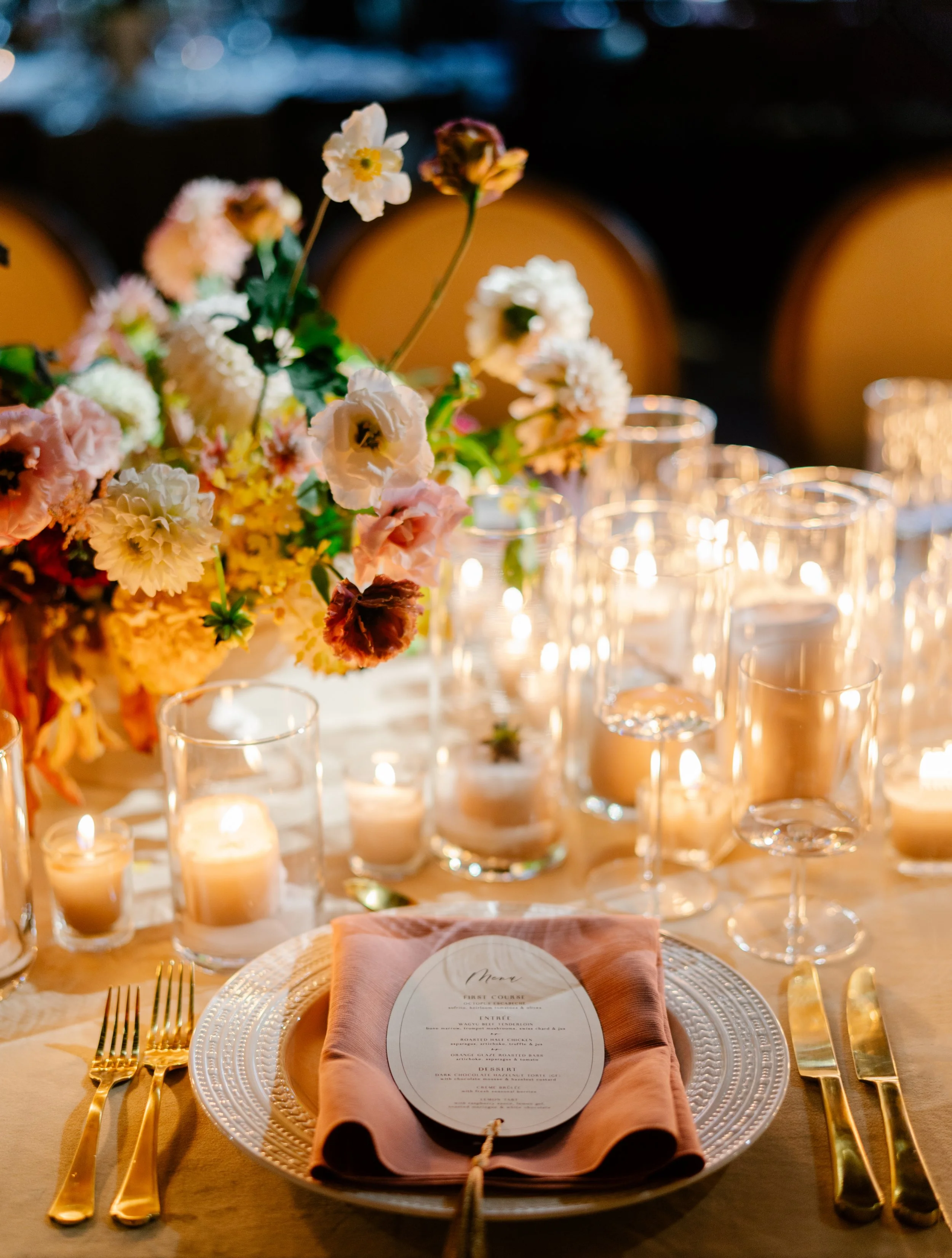 a floral arrangement with white, blush, and mauve florals surrounded by lit candles on a table