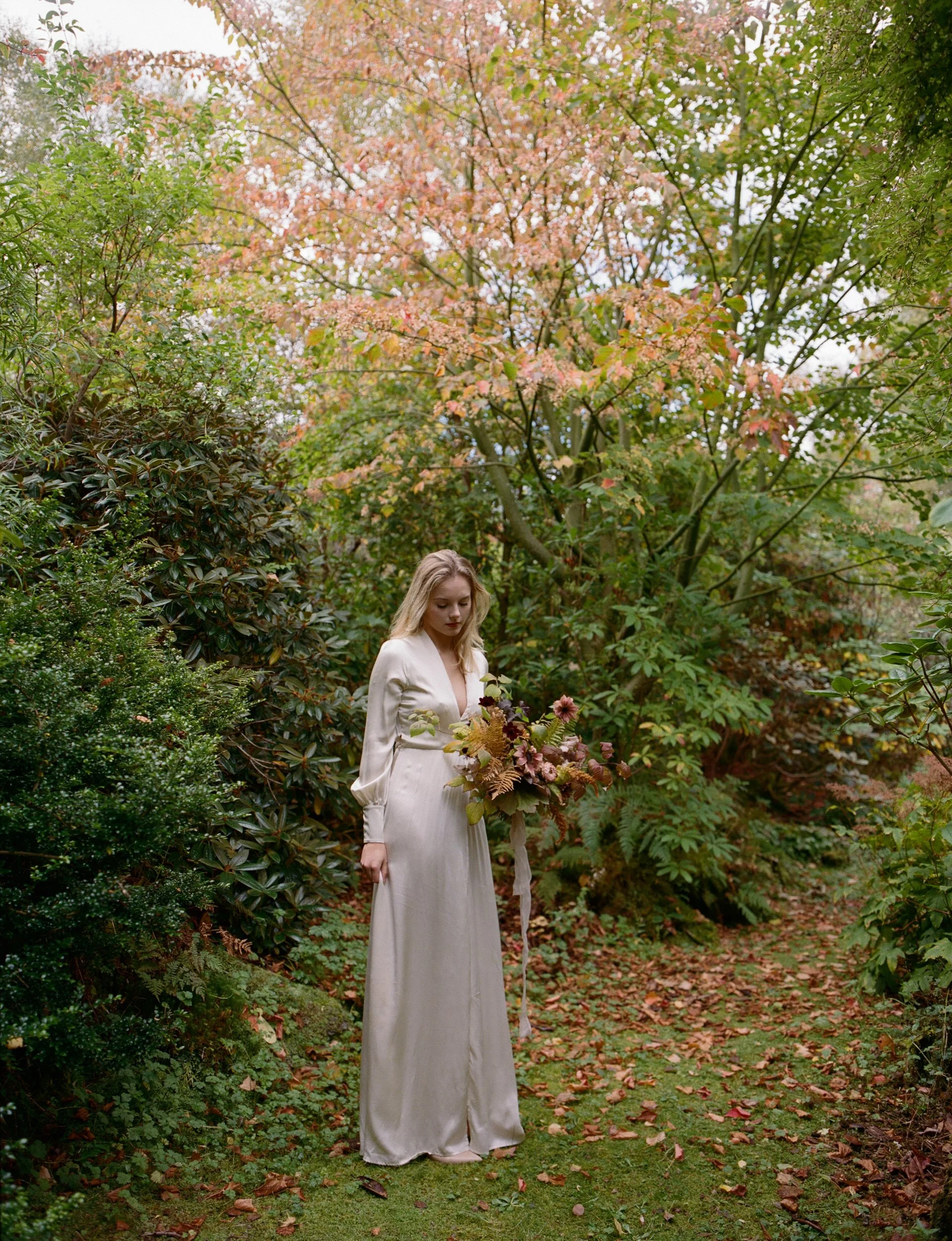 A woman in a white dress holding a bouquet of flowers stands in a lush garden with green foliage and autumn-colored trees.