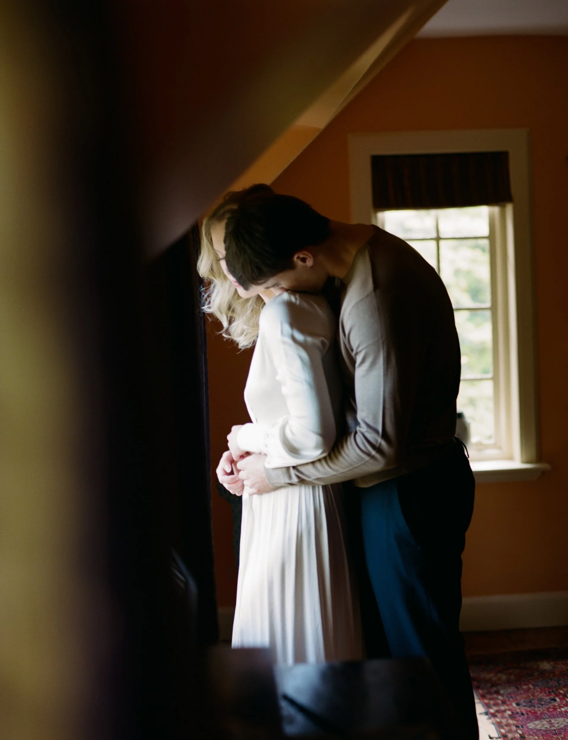 A couple embracing intimately indoors, with the man's head resting on the woman's shoulder, near a window with natural light coming in.