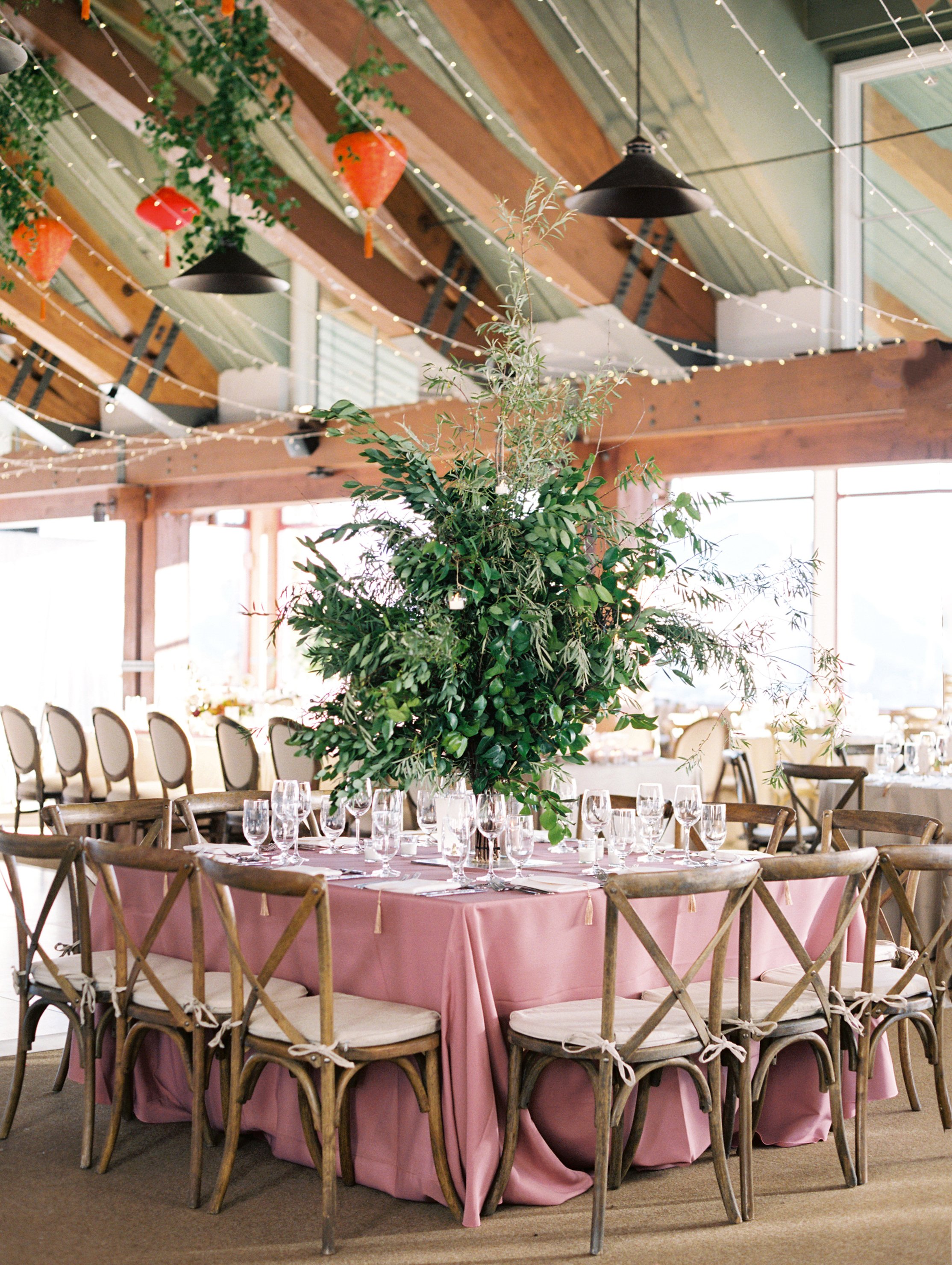 a tall leafy green arrangement on a table with a pink linen and wooden chairs around the table and hanging lights