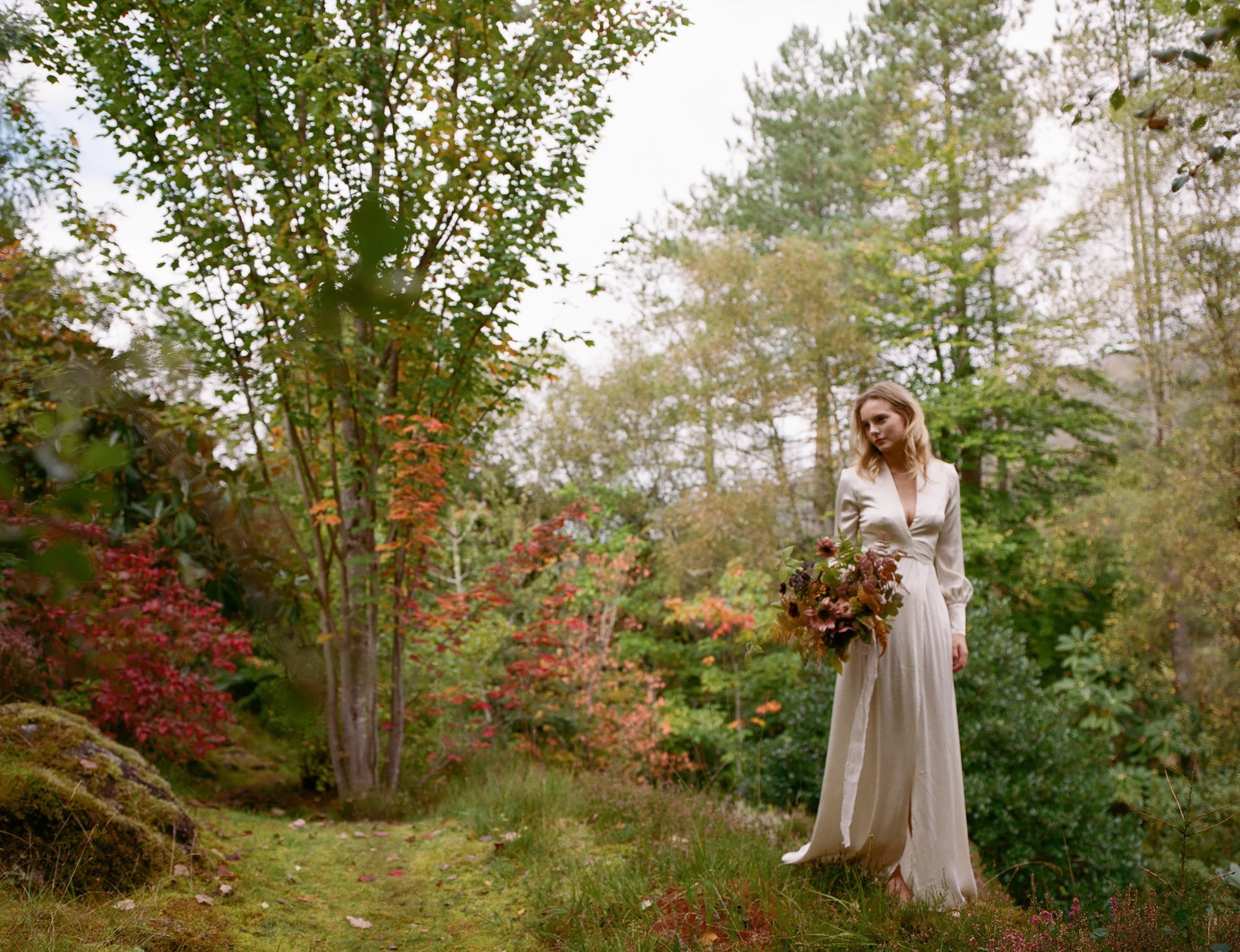 Woman in a white dress holding a bouquet of flowers stands outdoors on a grassy path surrounded by autumn trees.