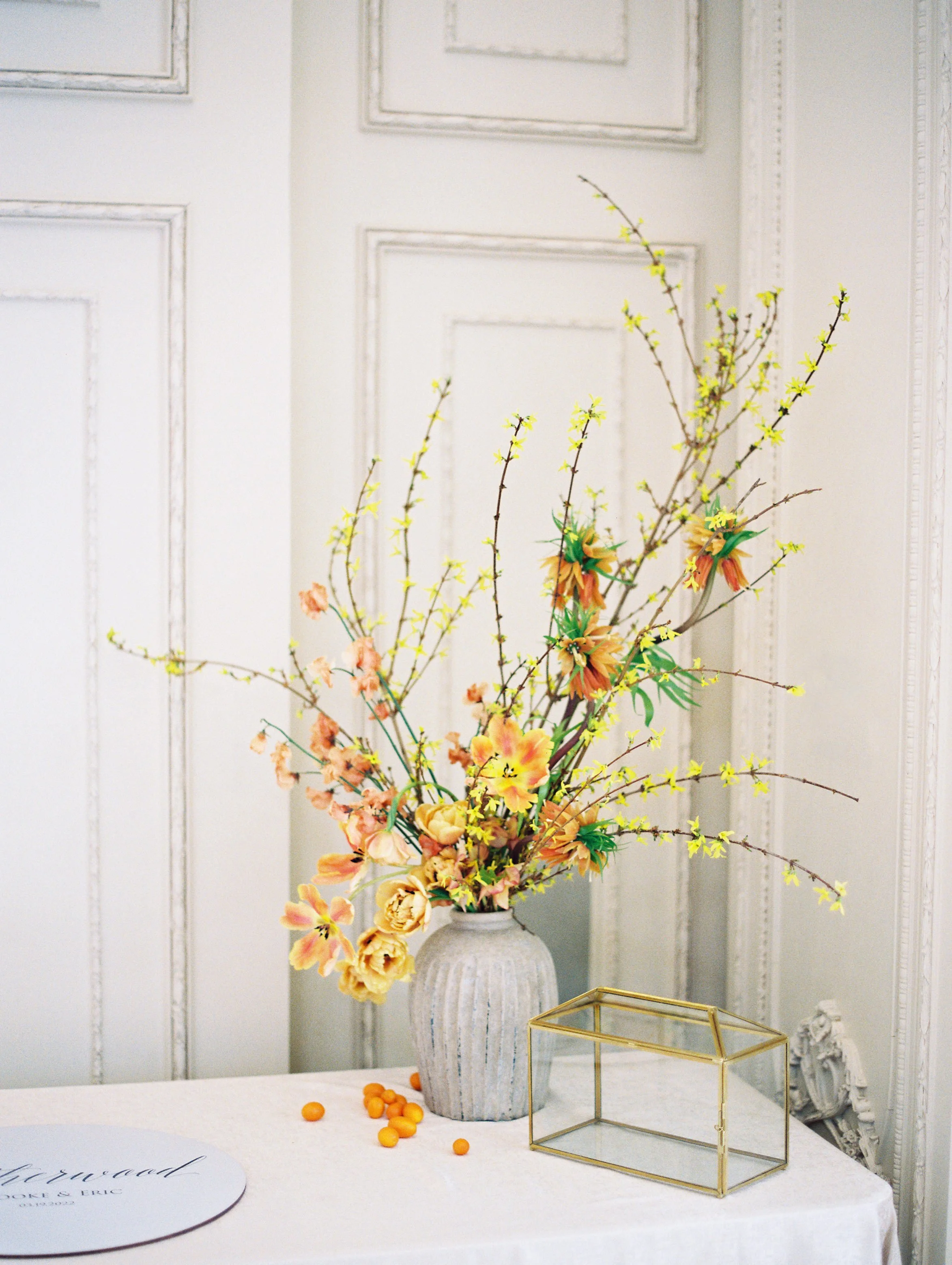 a floral arrangement with peach, yellow, and orange flowers in a grey vase with a gold lined glass box in a room