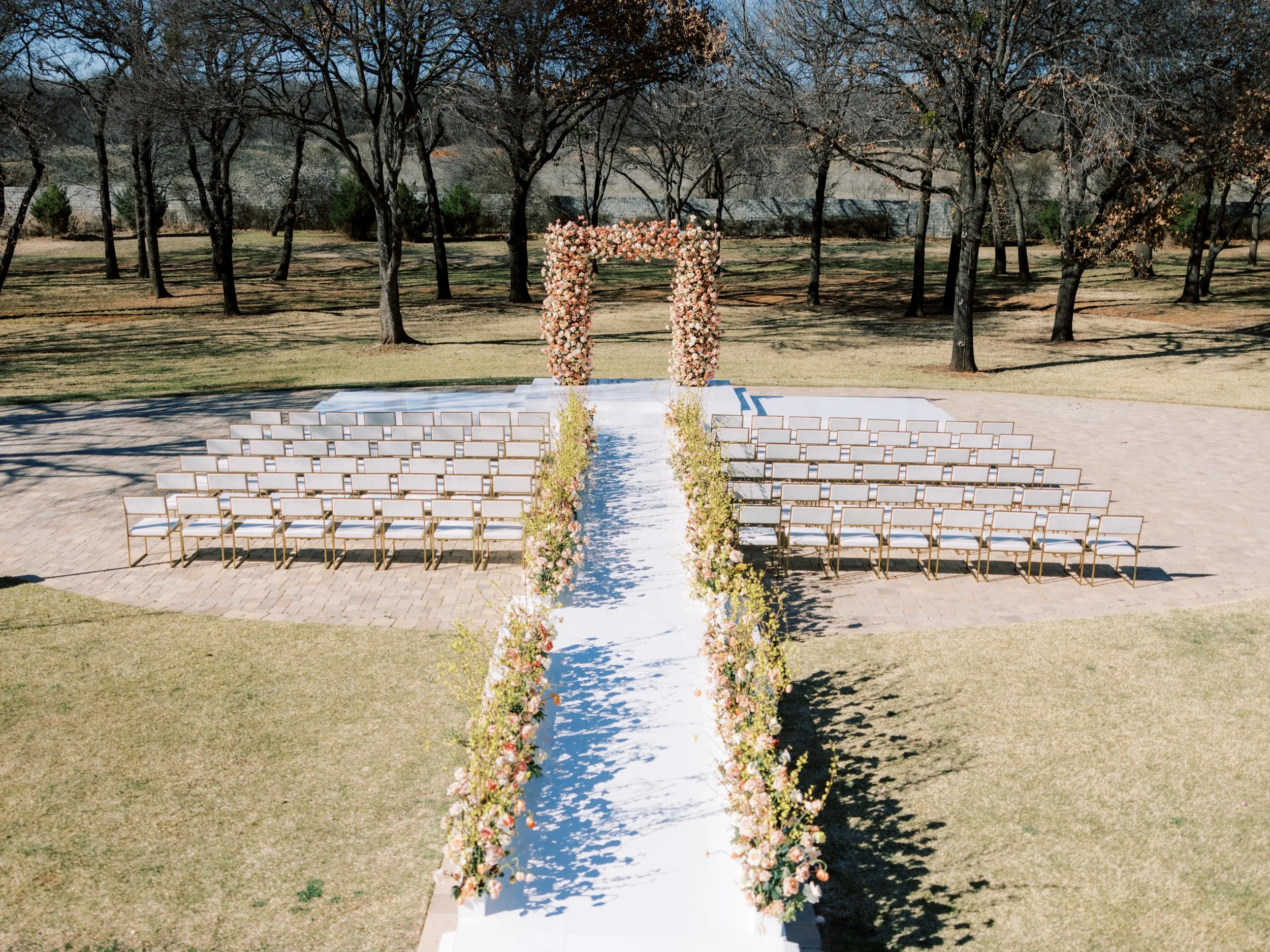 a large rectangle arch with peach and orange flowers and yellow flowers lining a white arch, all outside with bare trees in the background