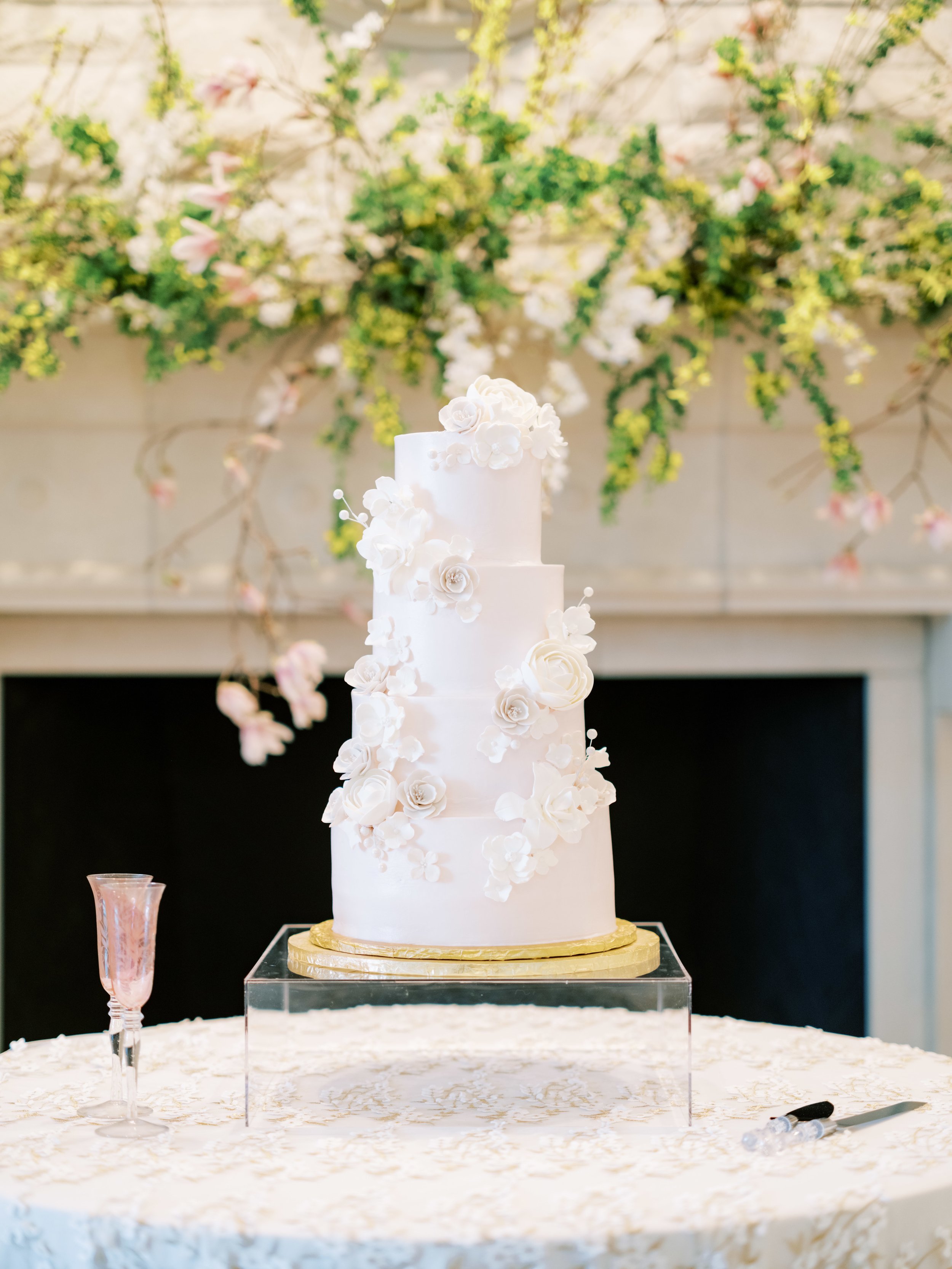 white tiered cake on acrylic box with fireplace behind it and yellow and spriggy flowers along it