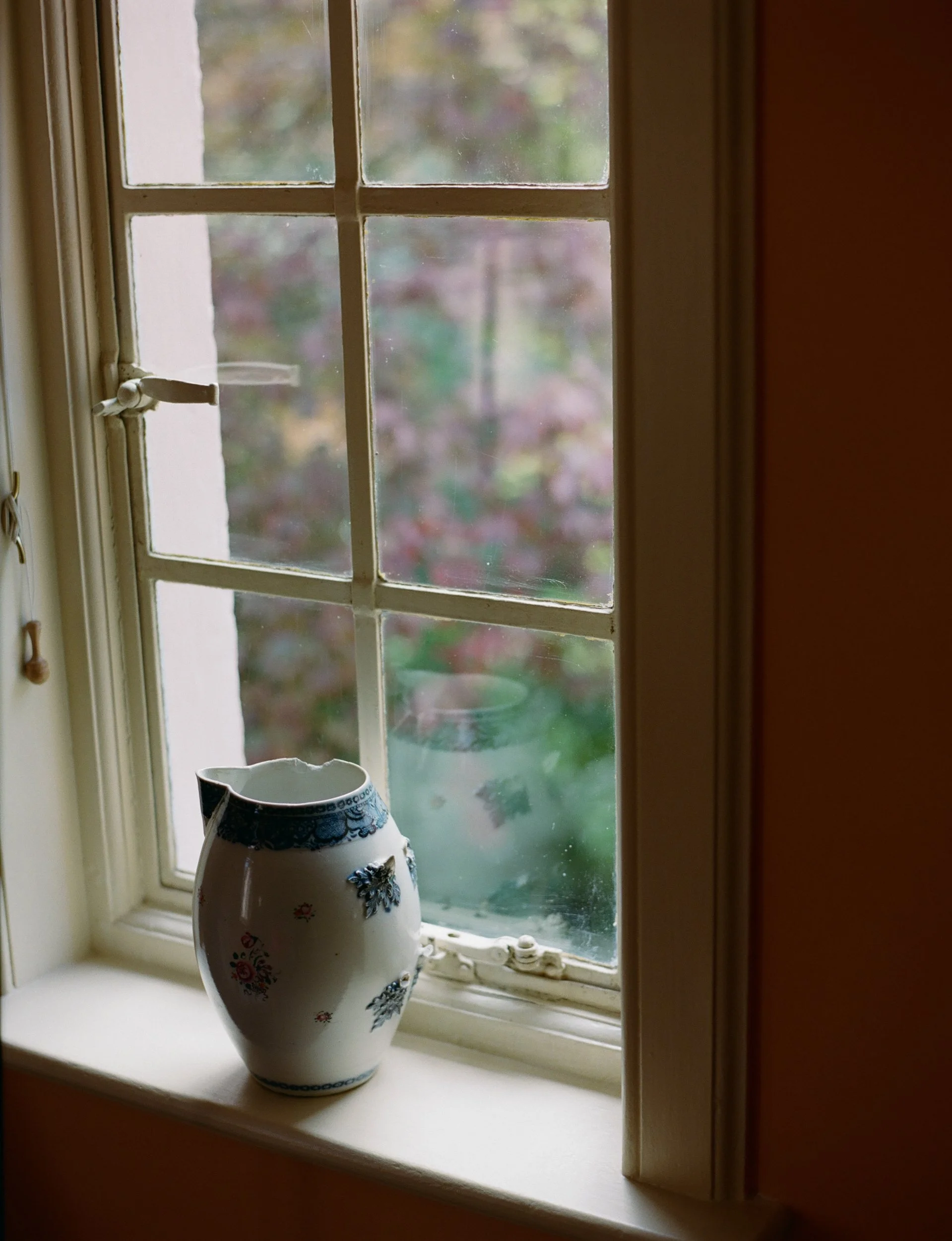 Decorative porcelain vase with floral patterns on a windowsill, with a blurred garden visible through the window.