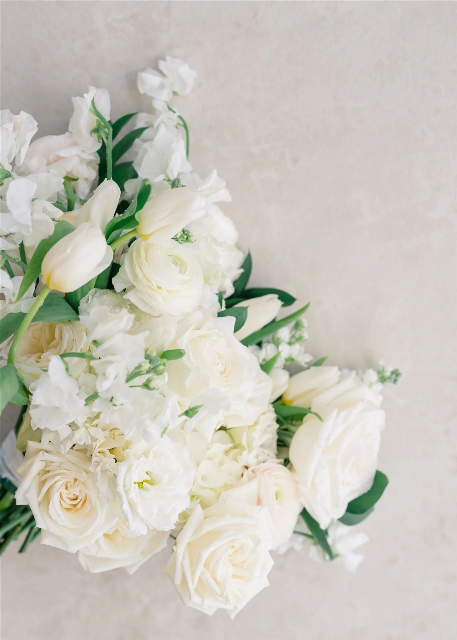 Bouquet of white flowers including roses, tulips, and other blossoms with green leaves on a light background.