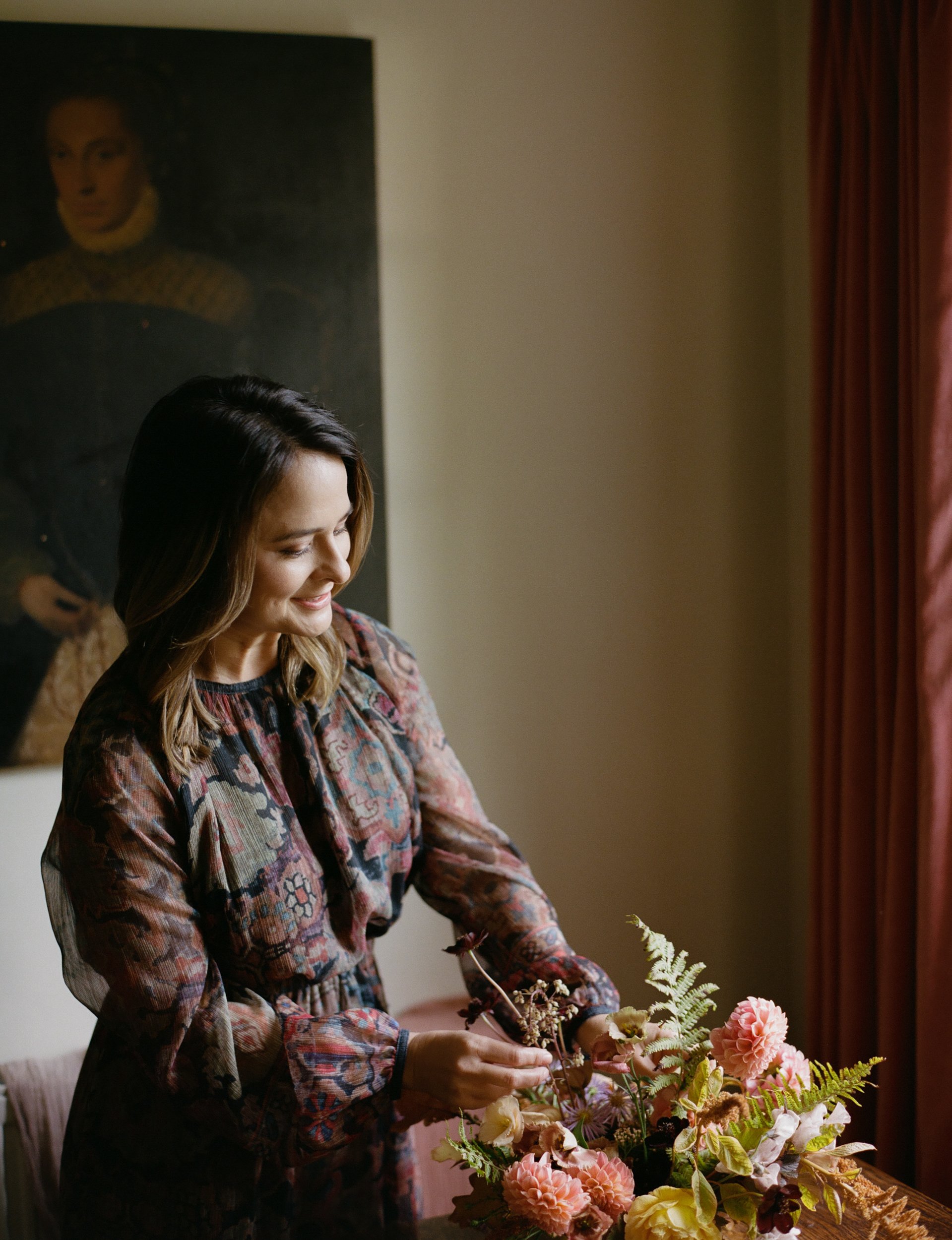 A woman arranging a flower bouquet indoors, with a painting on the wall and a window with red curtains in the background.
