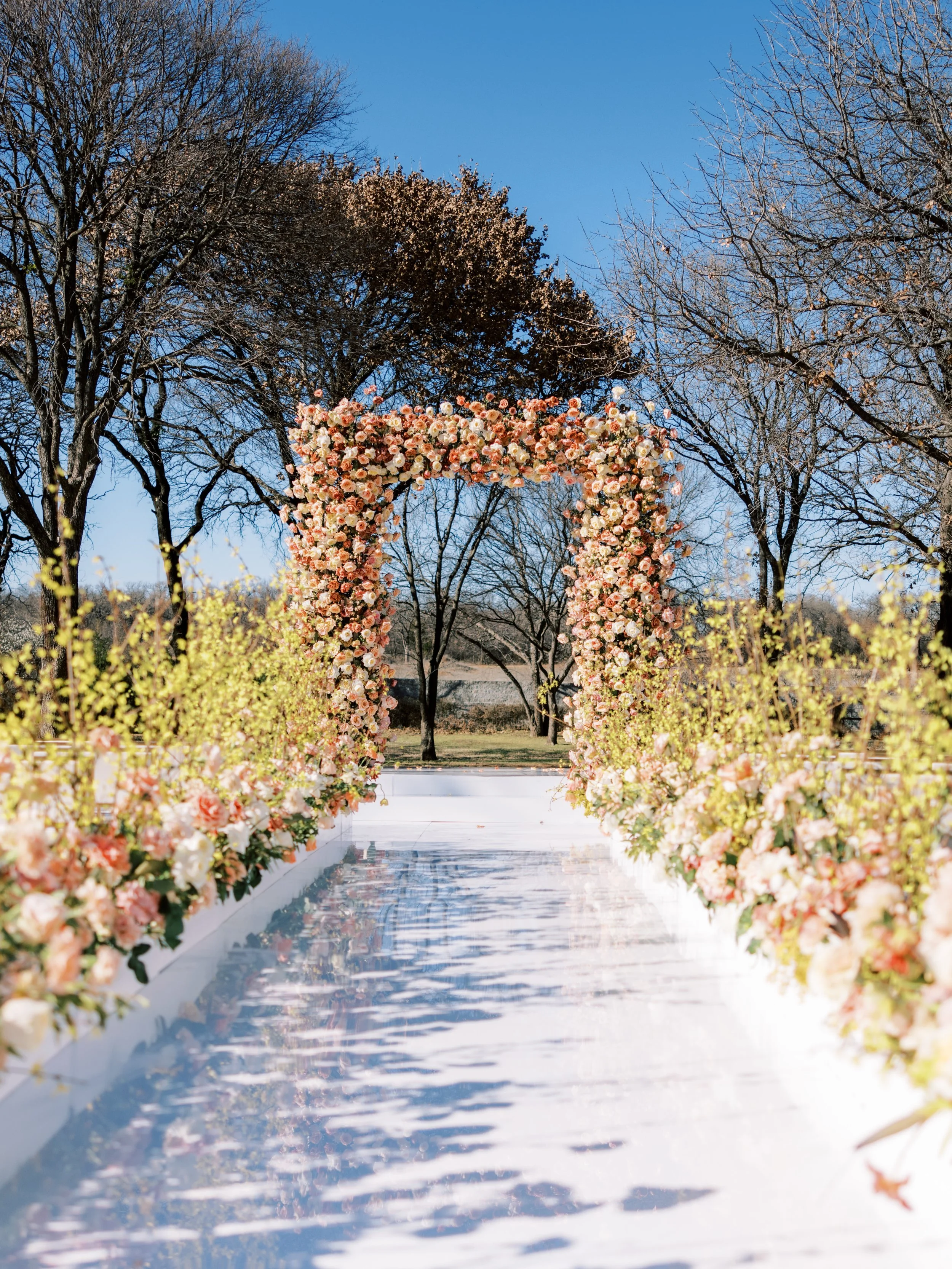 a white walkway leading up to a large rectangular arch filled with orange and peach flowers with yellow flowers lining the aisle