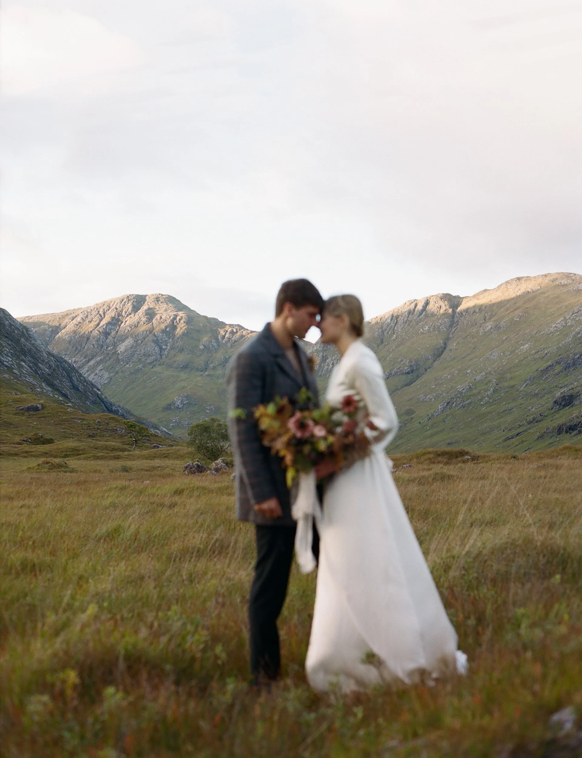 A couple dressed in wedding attire standing closely in a green field with mountains in the background, holding a bouquet of flowers, with their foreheads touching.