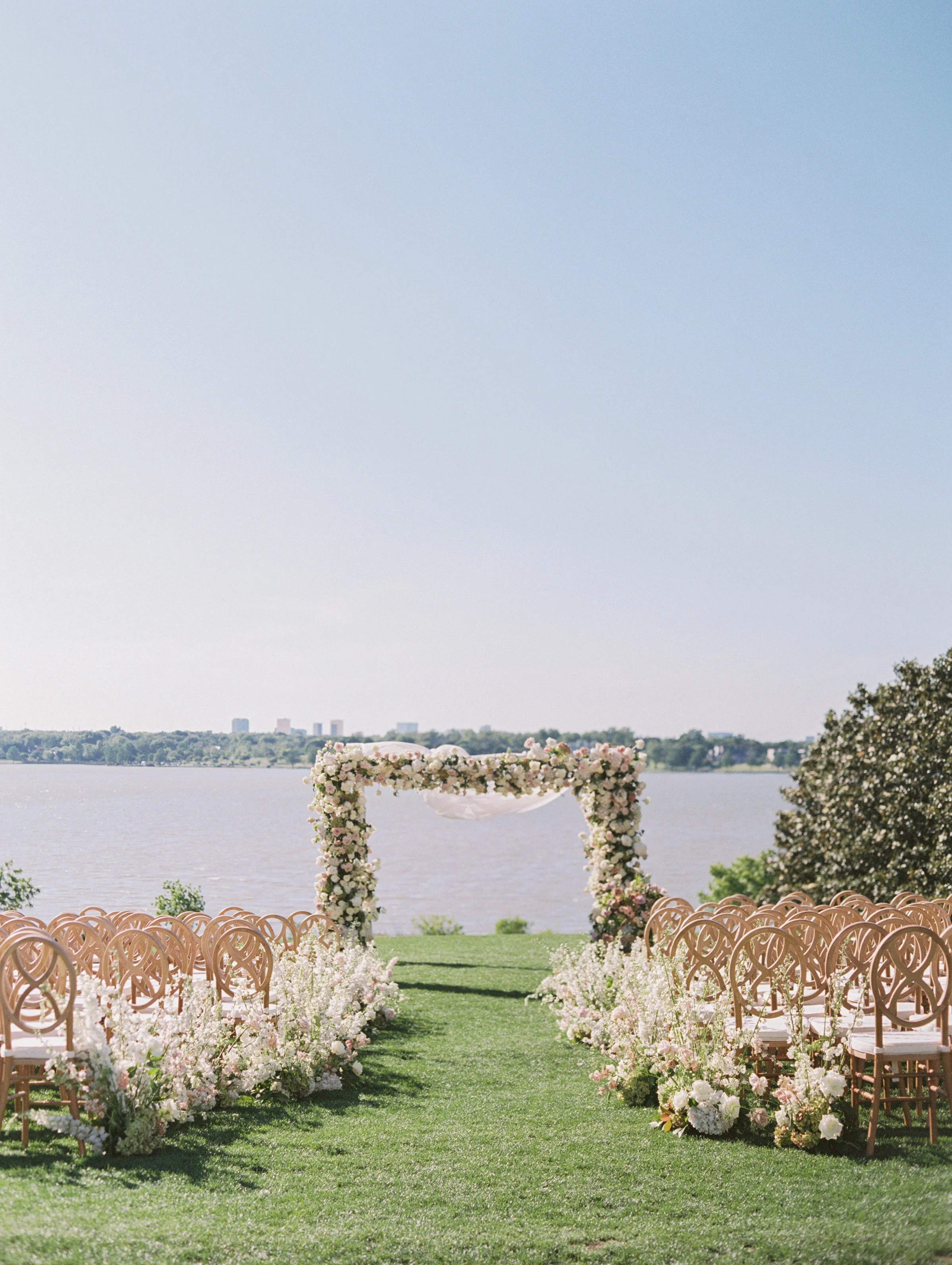 a chuppah with white and nude tall florals leading up to it with nude chairs with a lake in the background