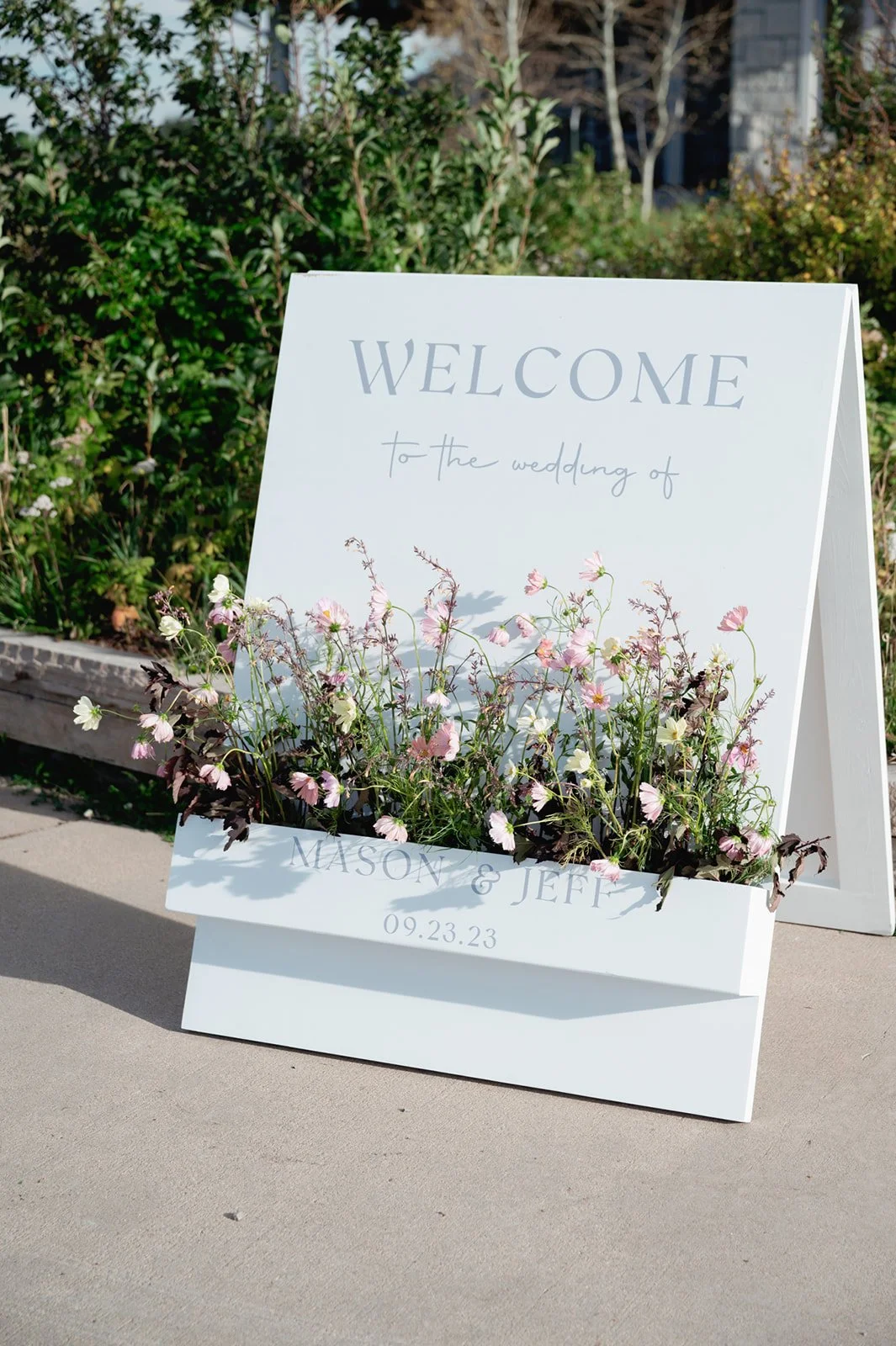 A white board welcome sign with florals growing out of it sitting outside