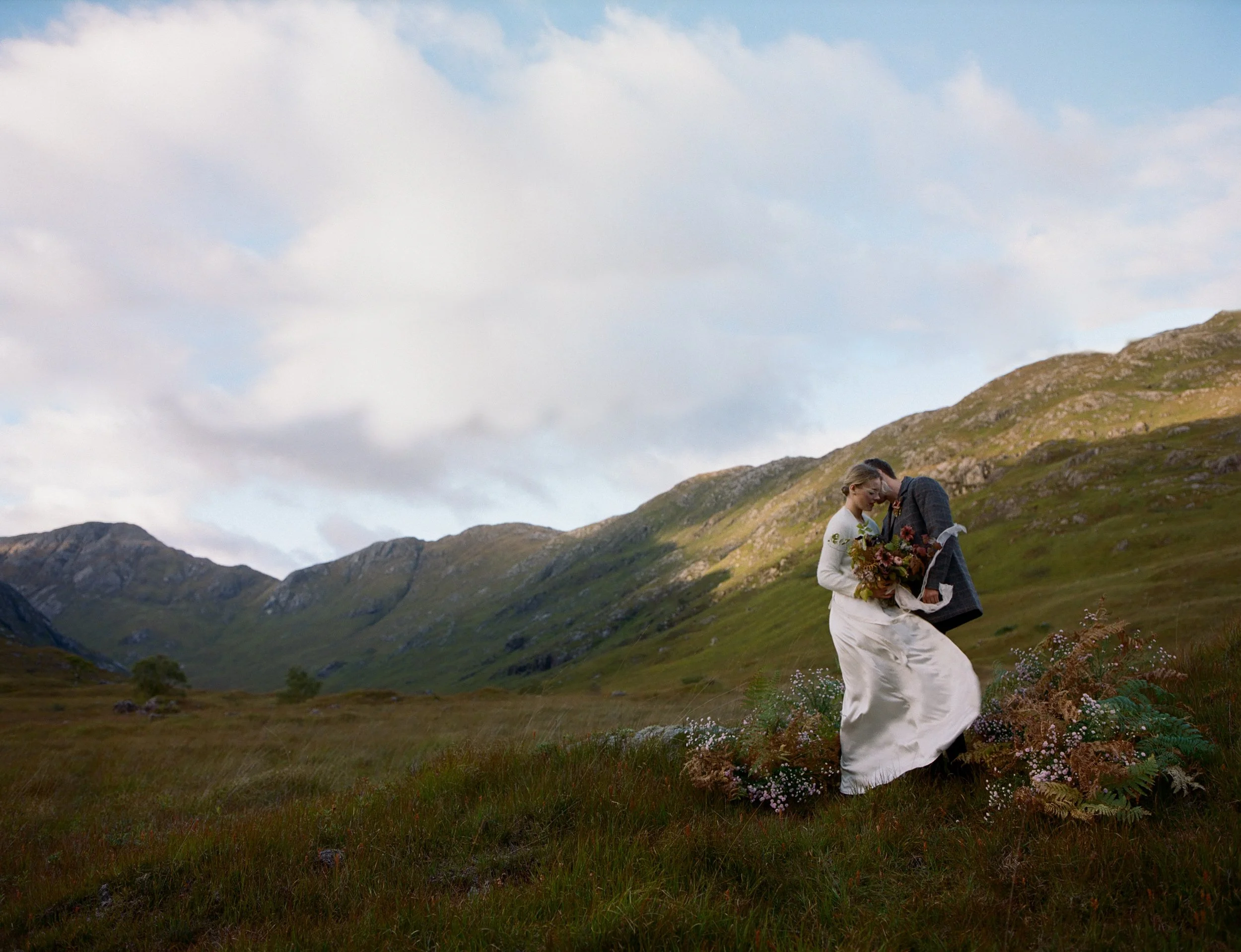 A couple dressed in wedding attire sharing a romantic moment outdoors in a scenic mountainous landscape, with wildflowers in the foreground and rolling green hills.
