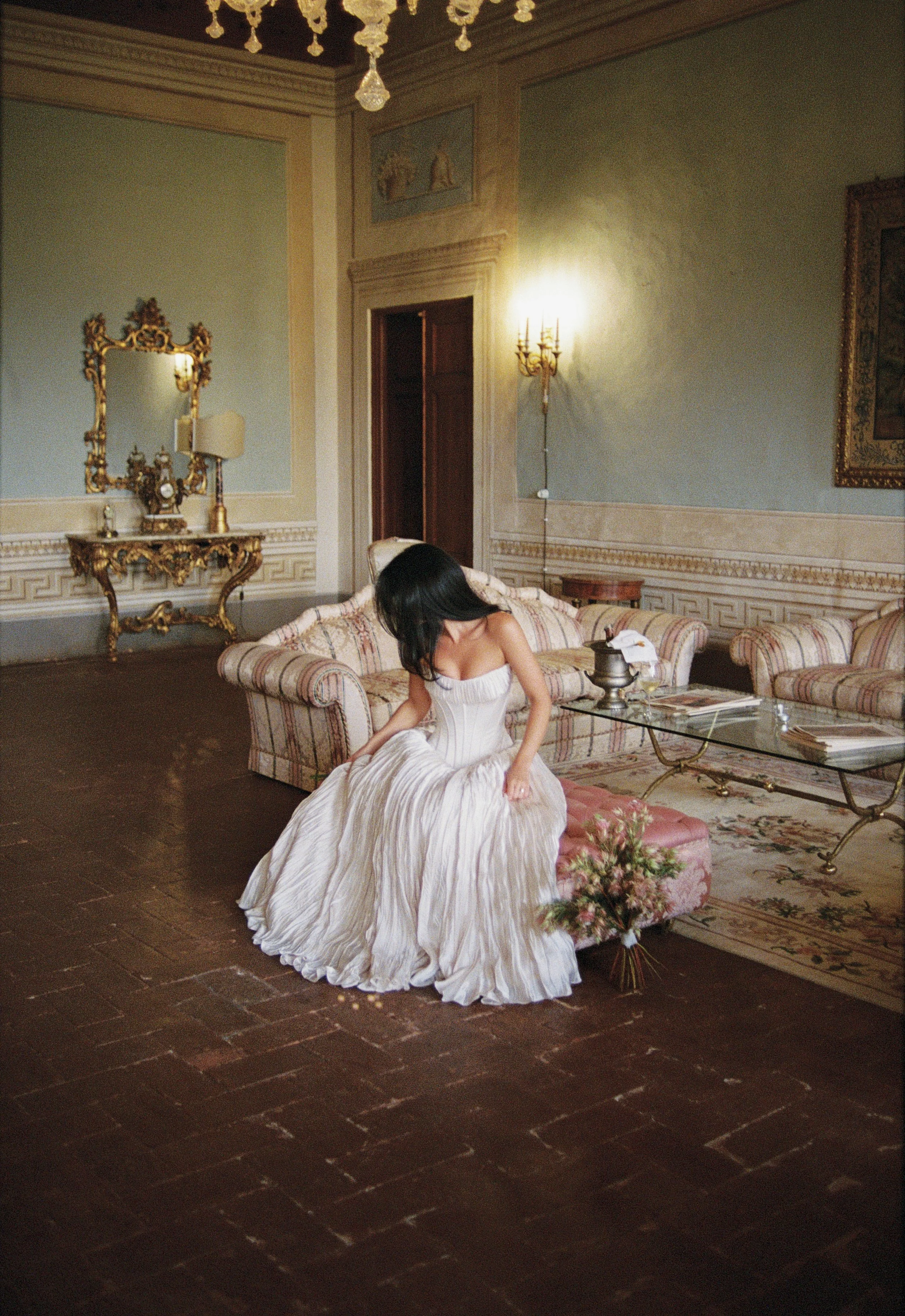 A  girl in a white dress is sitting on a chair with a bouquet of flowers next to her. She is looking down. Pale blue walls