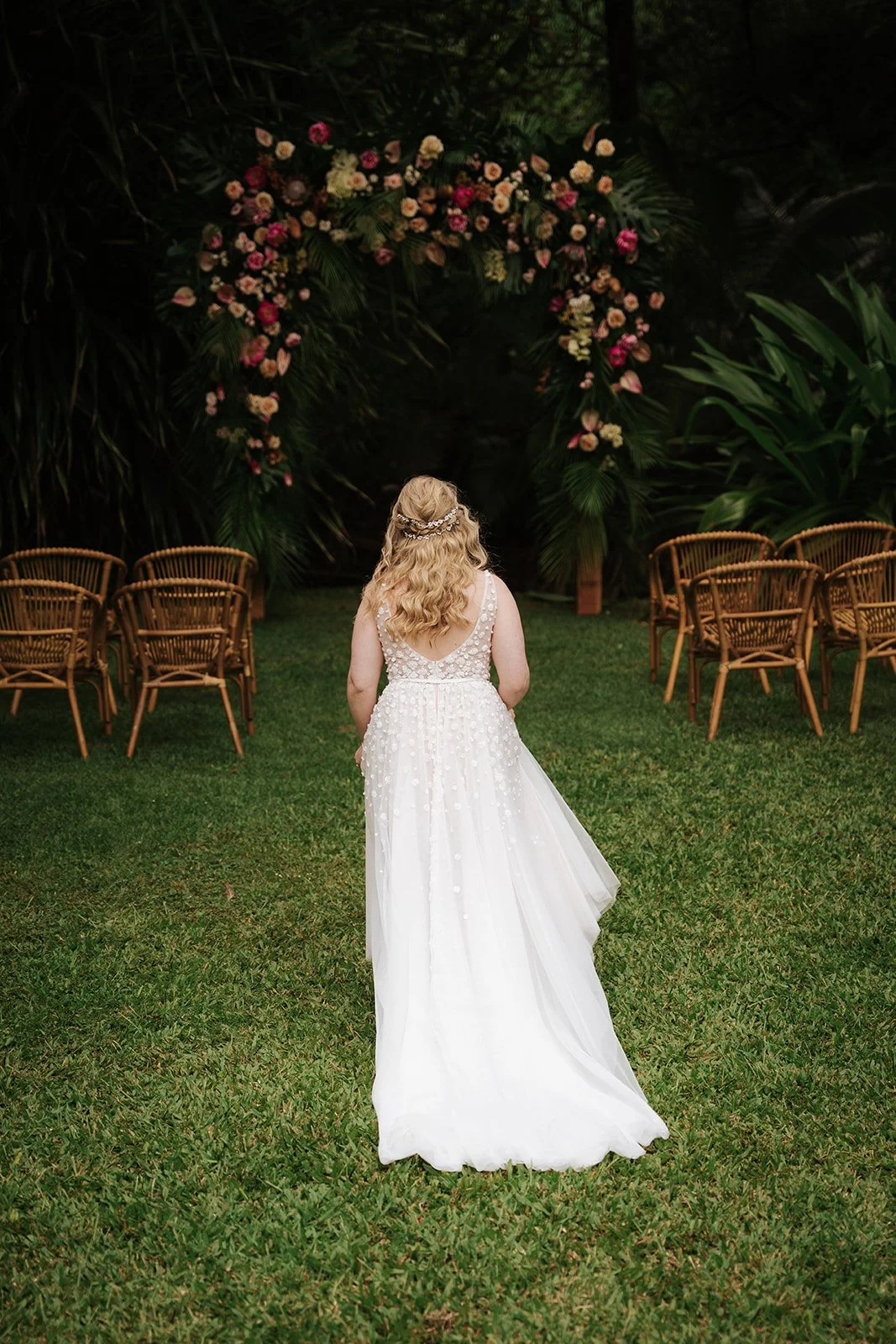 A bride in a white dress is walking to a floral arch