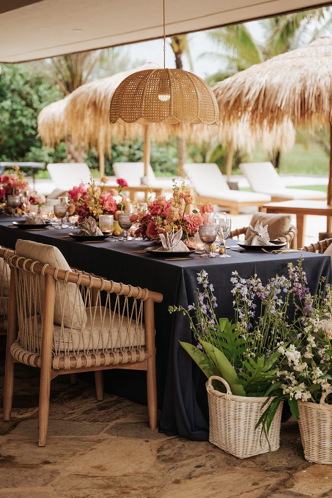 table with blue linen and wood and wicker chairs with a basket of flowers and pink arrangements on table and straw canopies