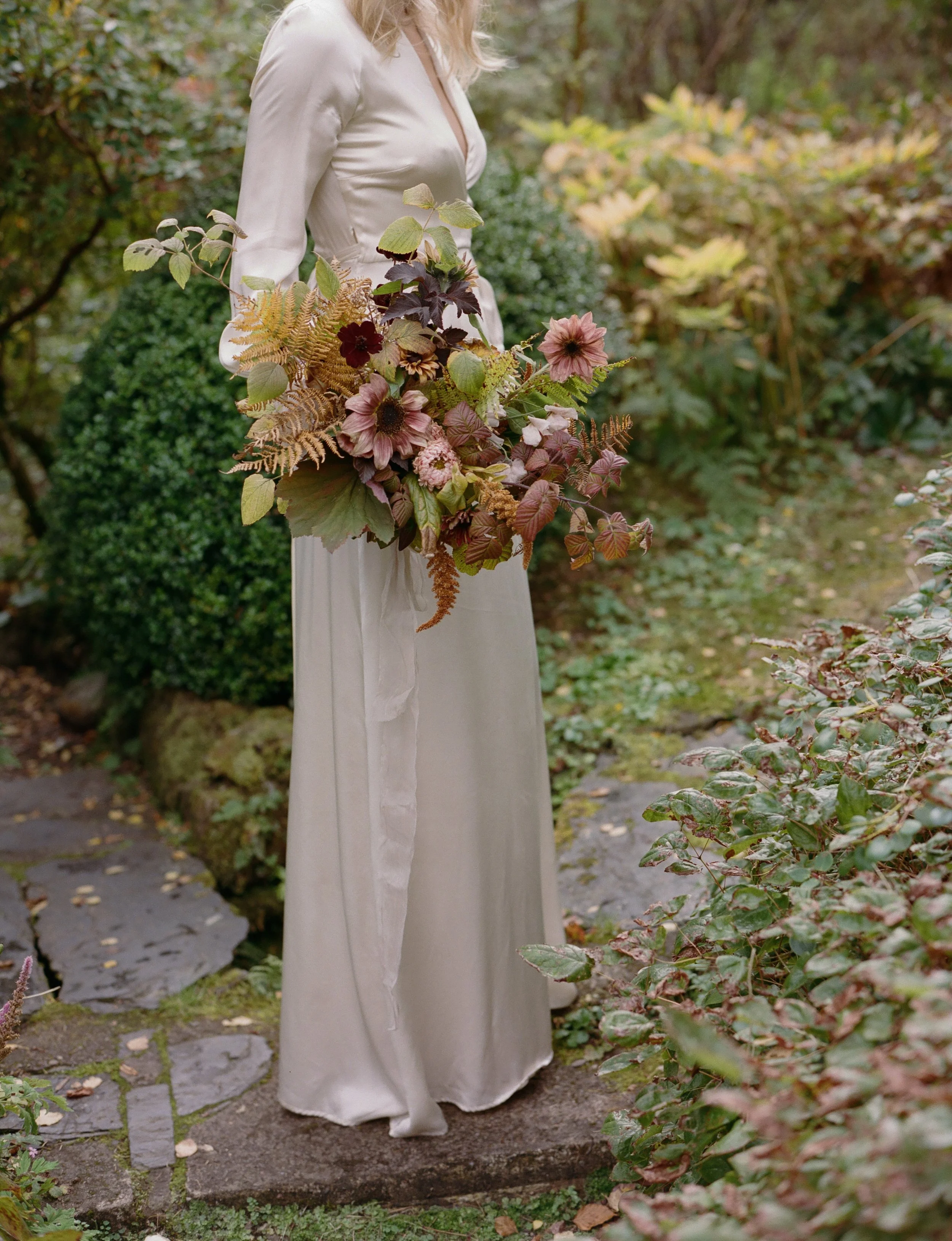A person in an elegant white dress standing outdoors on a stone pathway, holding a large bouquet of pink, purple, and green foliage surrounded by greenery.