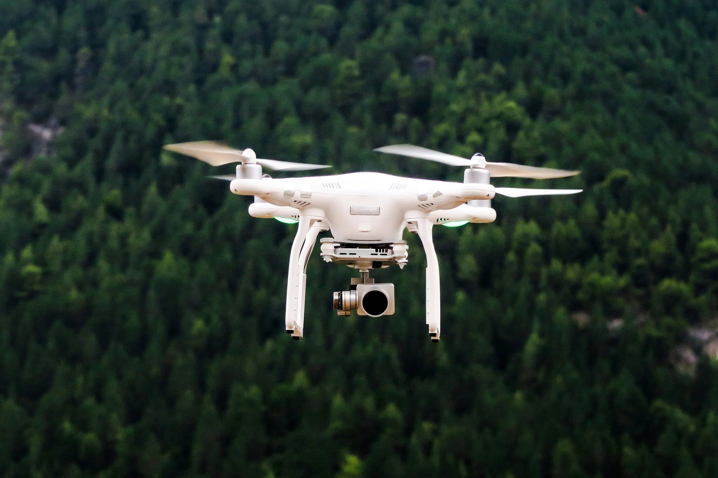 Drone blanc avec caméra suspendue en vol devant une forêt dense.