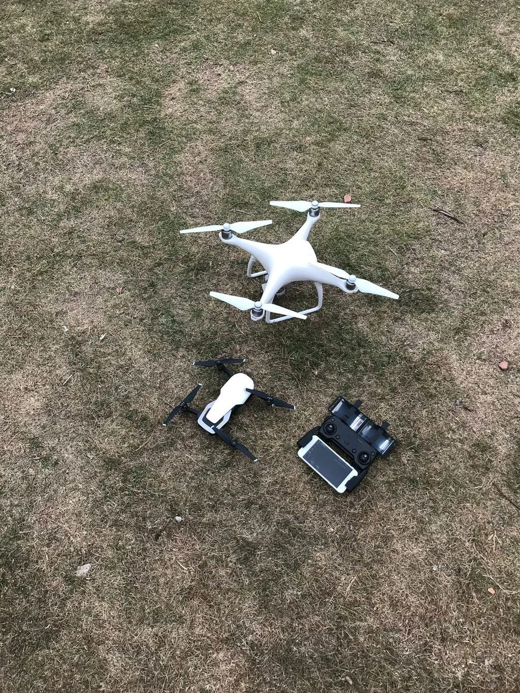Un drone blanc avec quatre hélices, un drone noir avec caméra, une télécommande et une tablette sur un sol en herbe.