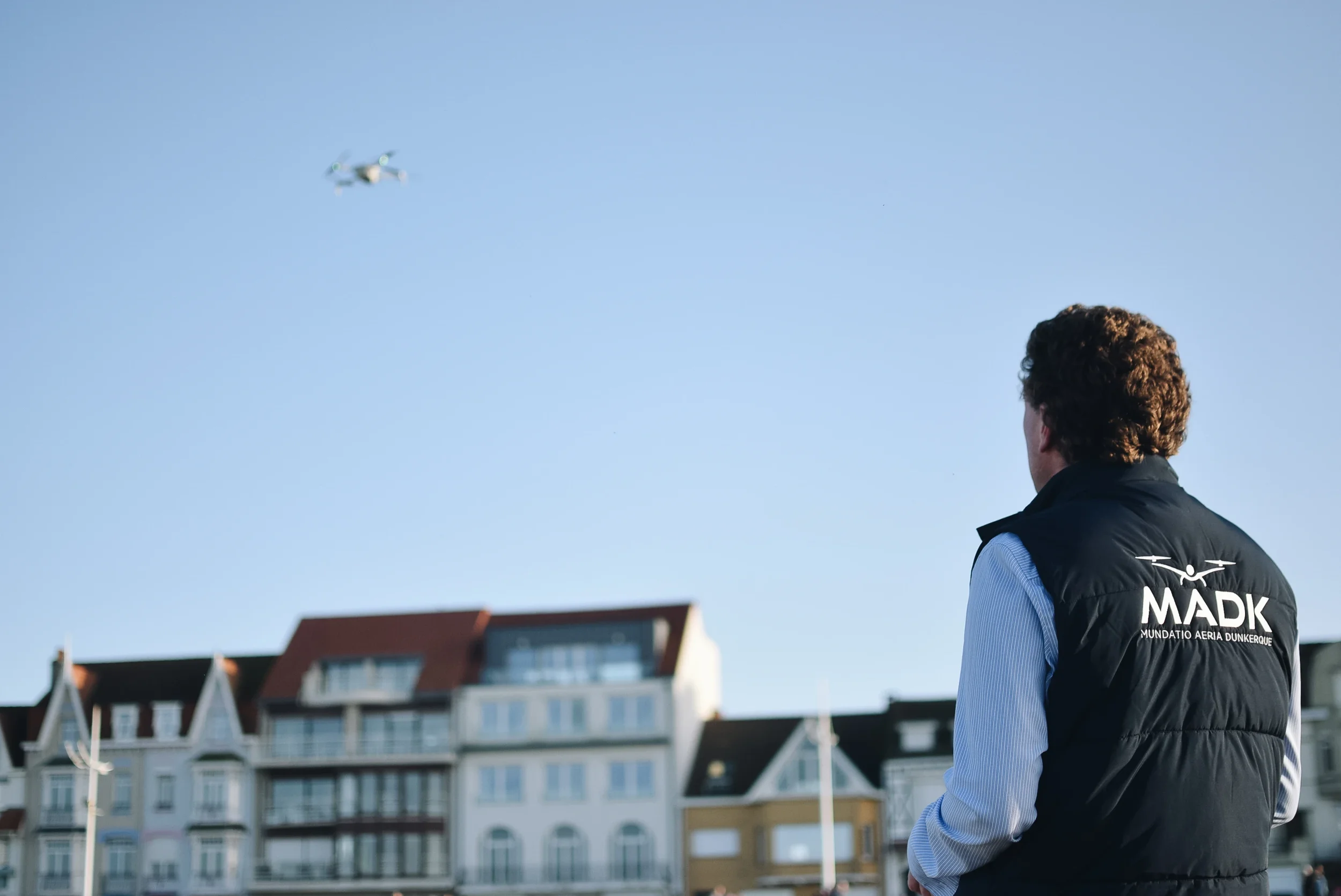 Une personne portant une veste avec le logo MADK pilote un drone dans le ciel au-dessus de bâtiments résidentiels.