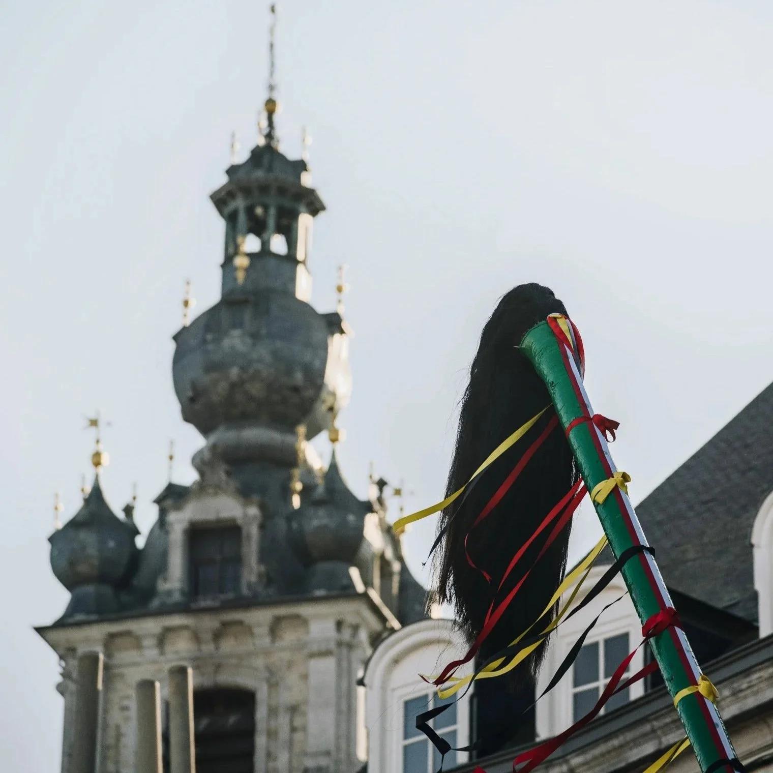 Un chandelier de fête avec des rubans multicolores attachés, placé devant un bâtiment historique avec une tour à bulbe en arrière-plan.