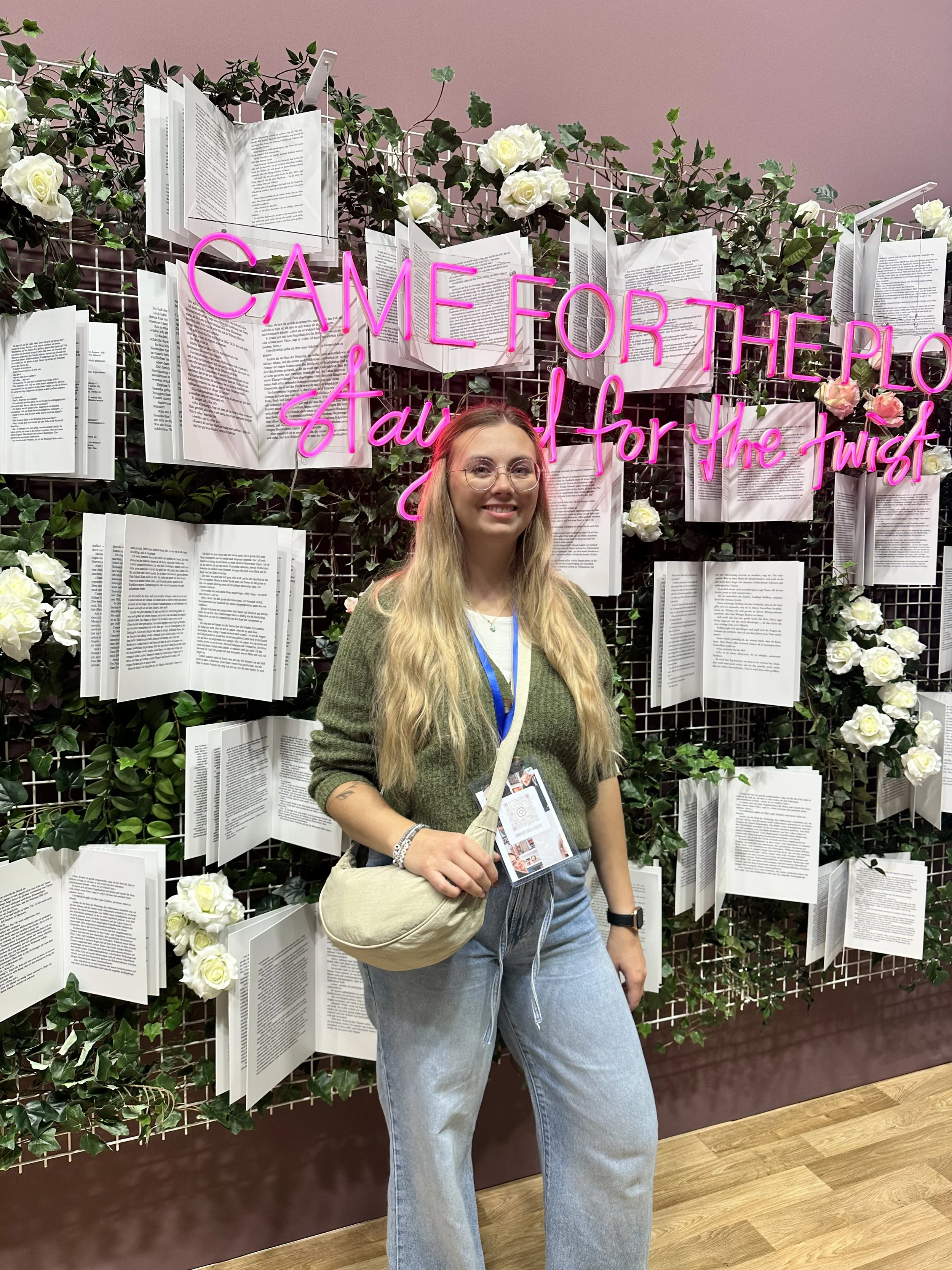 Lissa Göris steht vor einem Fotospot mit neon pinken Schriftzug und aufgeklappten Büchern vor grünem Hintergrund. Der Schriftzug lautet "Came for the Plot, stay for the Twist."