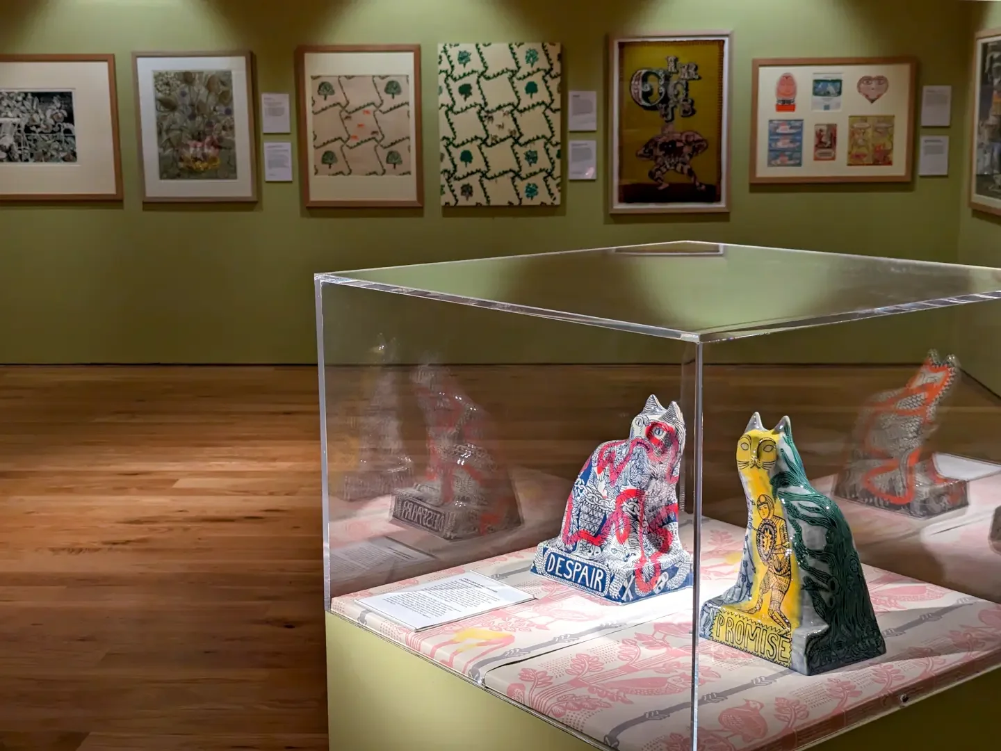 Art exhibit in a gallery with paintings on the wall and a glass display case with colourful ceramic cat sculptures.