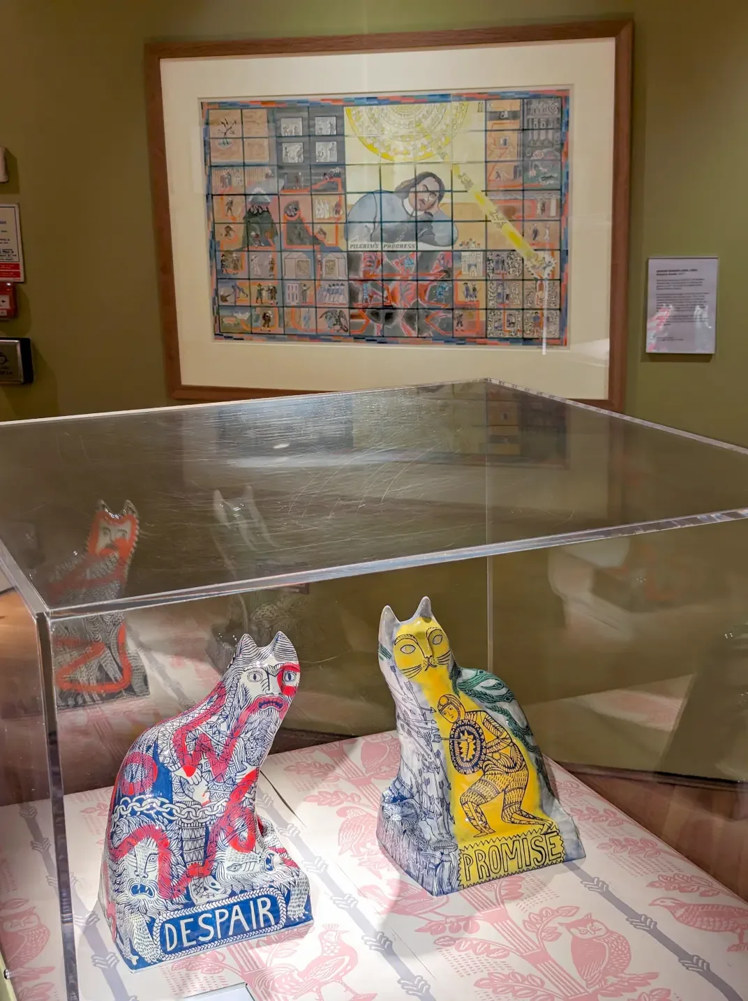 Colorful art display with two decorated cat sculptures labeled "Despair" and "Promise" inside a glass case, with a framed artwork on the wall depicting a person surrounded by squares and symbols in the background.