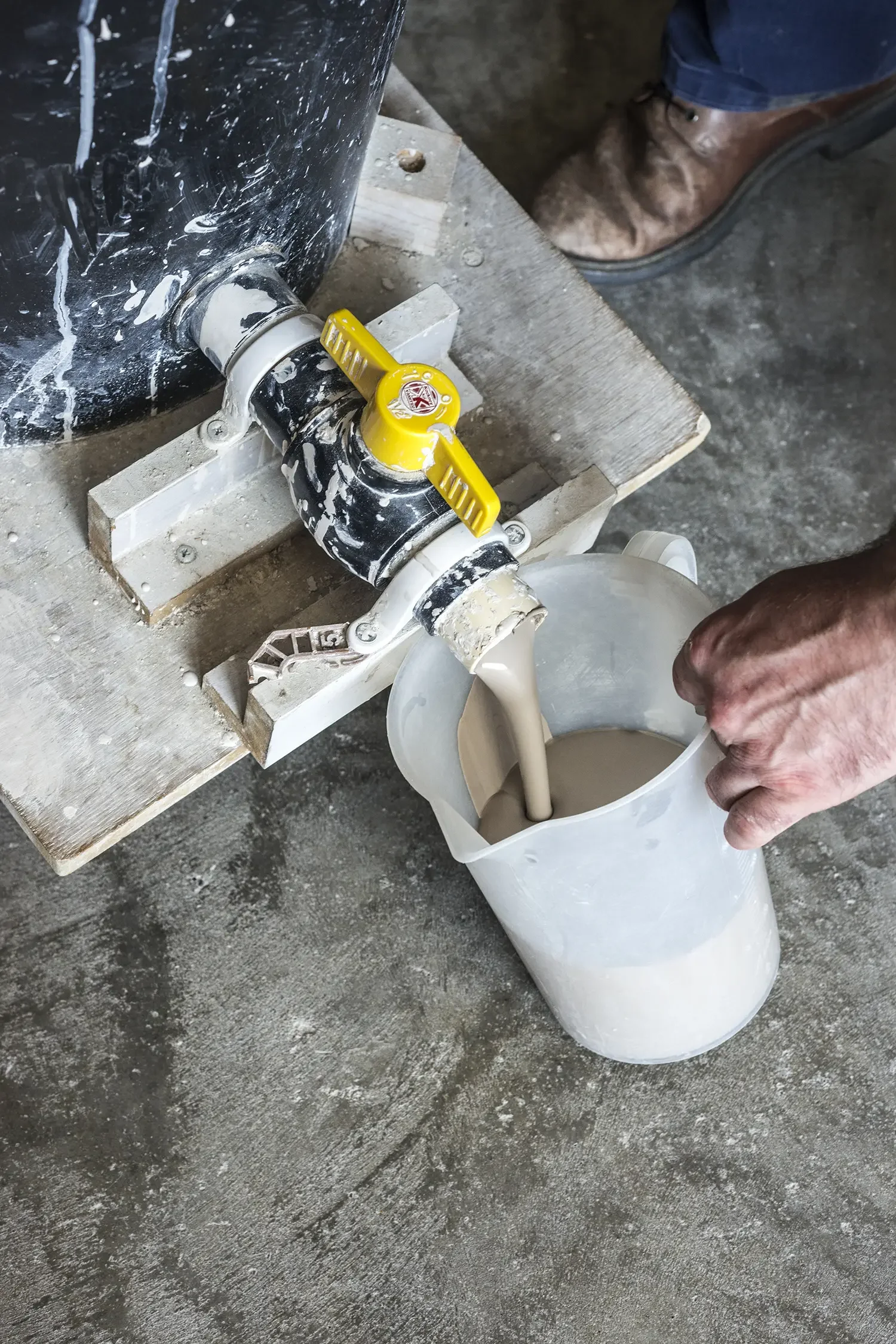 Bill Brookes pouring clay slip into a jug from a tap connected to a black container. The scene is on a concrete floor.