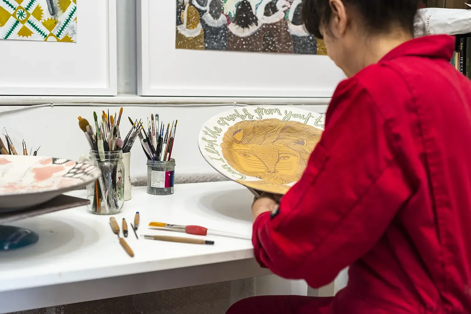 Vicky Lindo in a red overalls is working on a lion illustration on a ceramic plate, surrounded by various tools for sgraffito decorating.