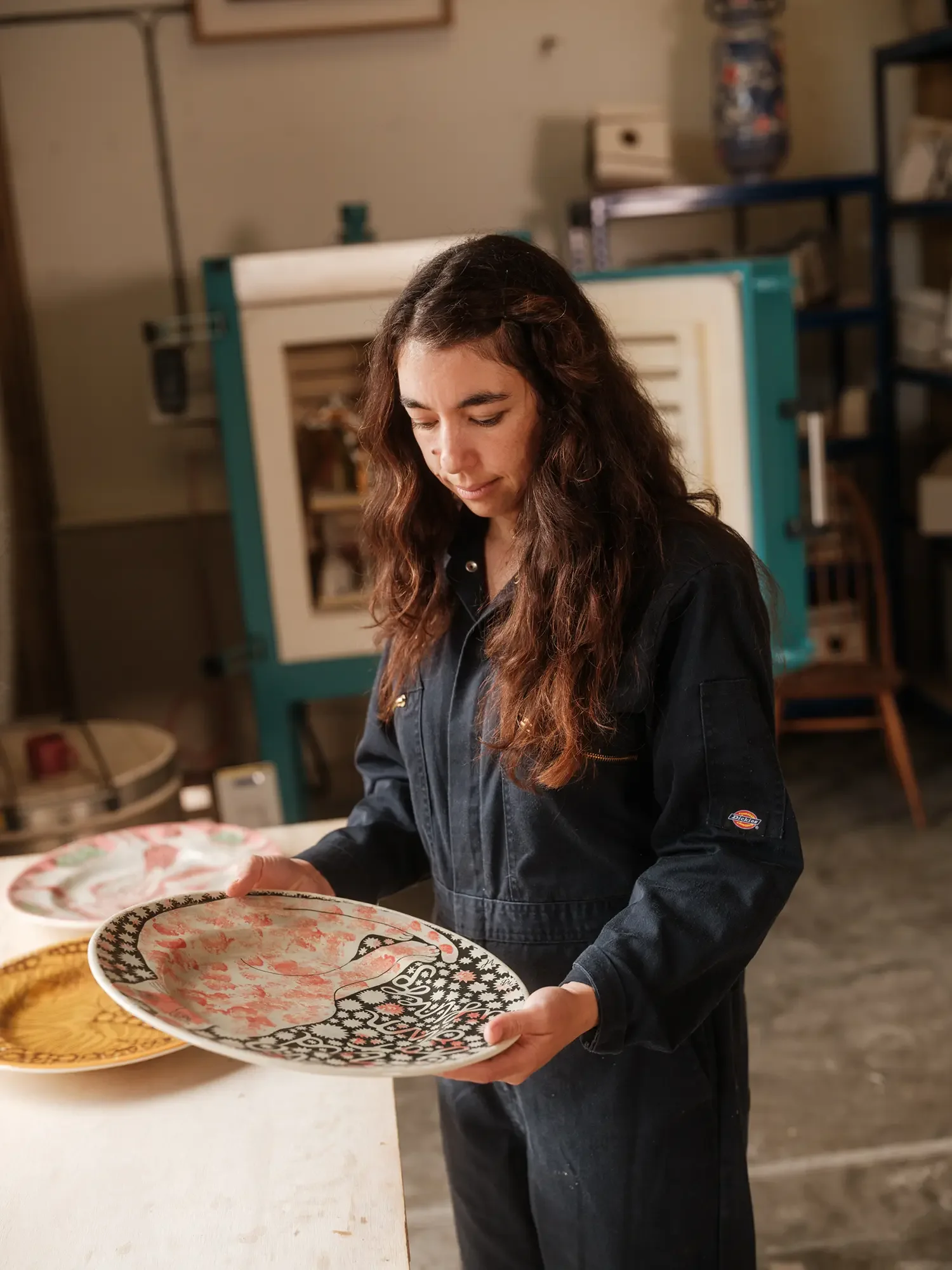 Vicky Lindo with long brown curly hair wearing a black overalls looking down at ceramic plates with various patterns and colours on a white surface in a workshop or studio.