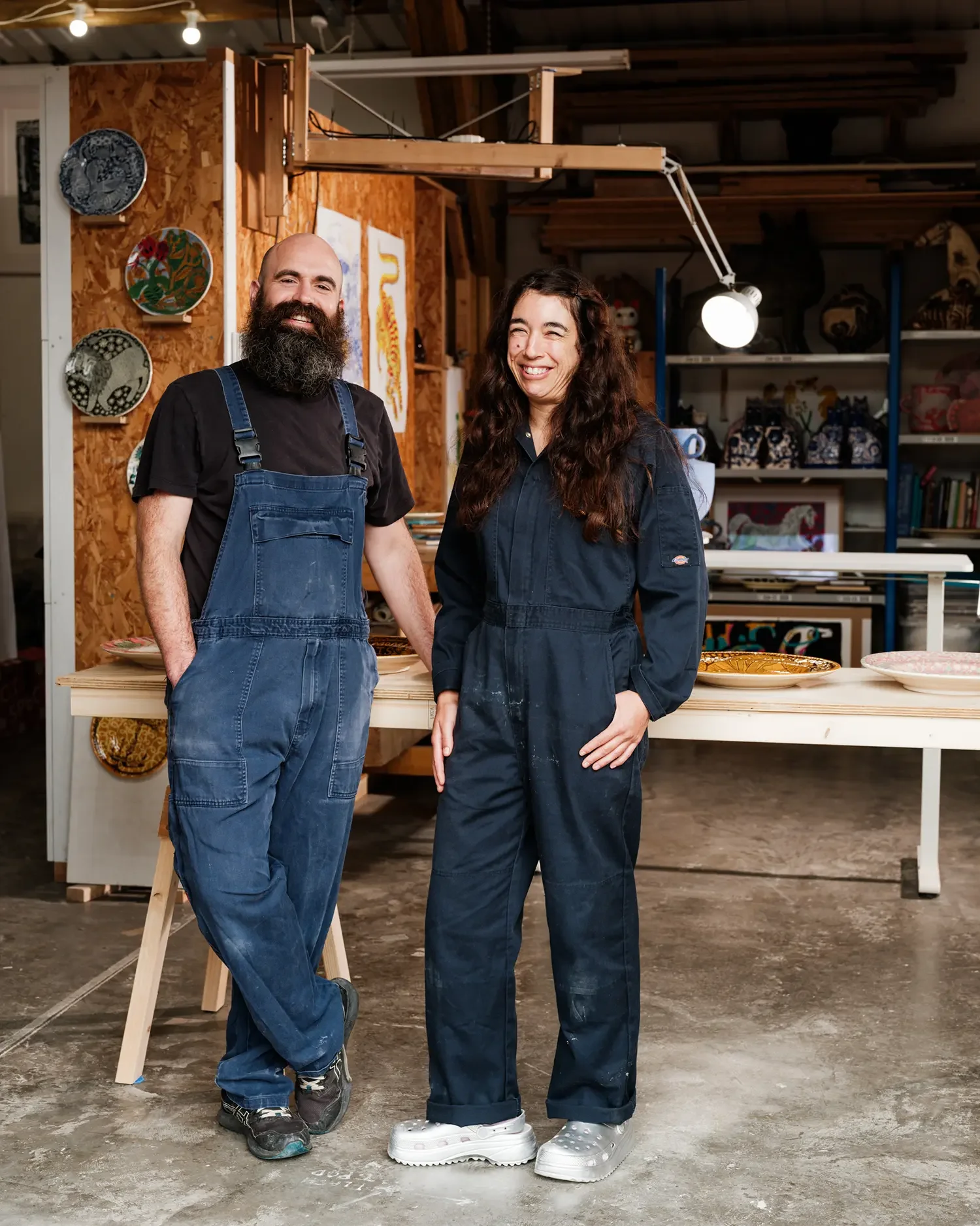 Bill Brookes and Vicky Lindo in work overalls smiling in an art studio with finished ceramics on the wall and art supplies on shelves in the background.