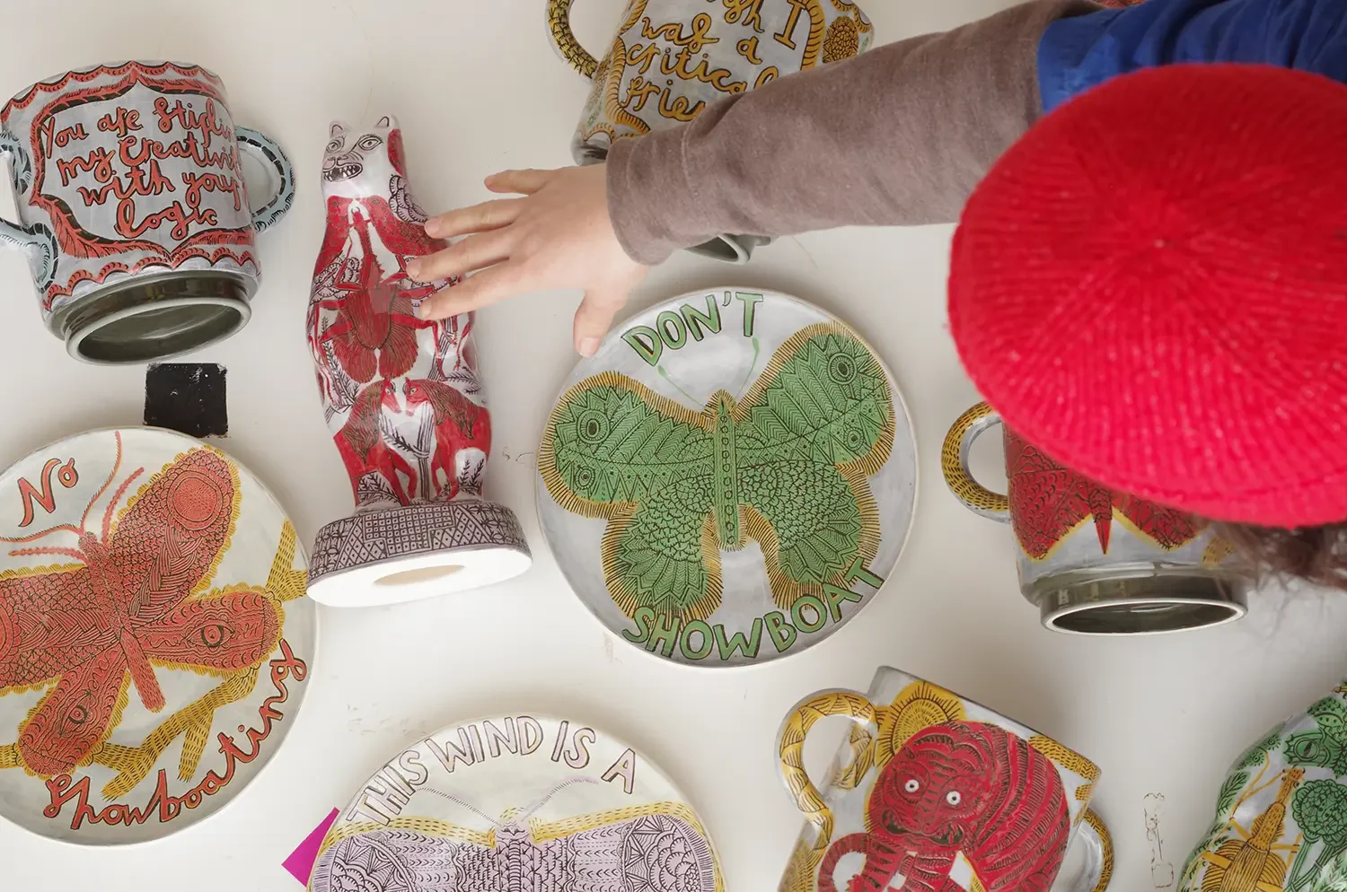 Decorative plates and vessels with colourful butterfly, bear, and other patterns arranged on a white surface, with a hand reaching towards a butterfly plate in the centre.