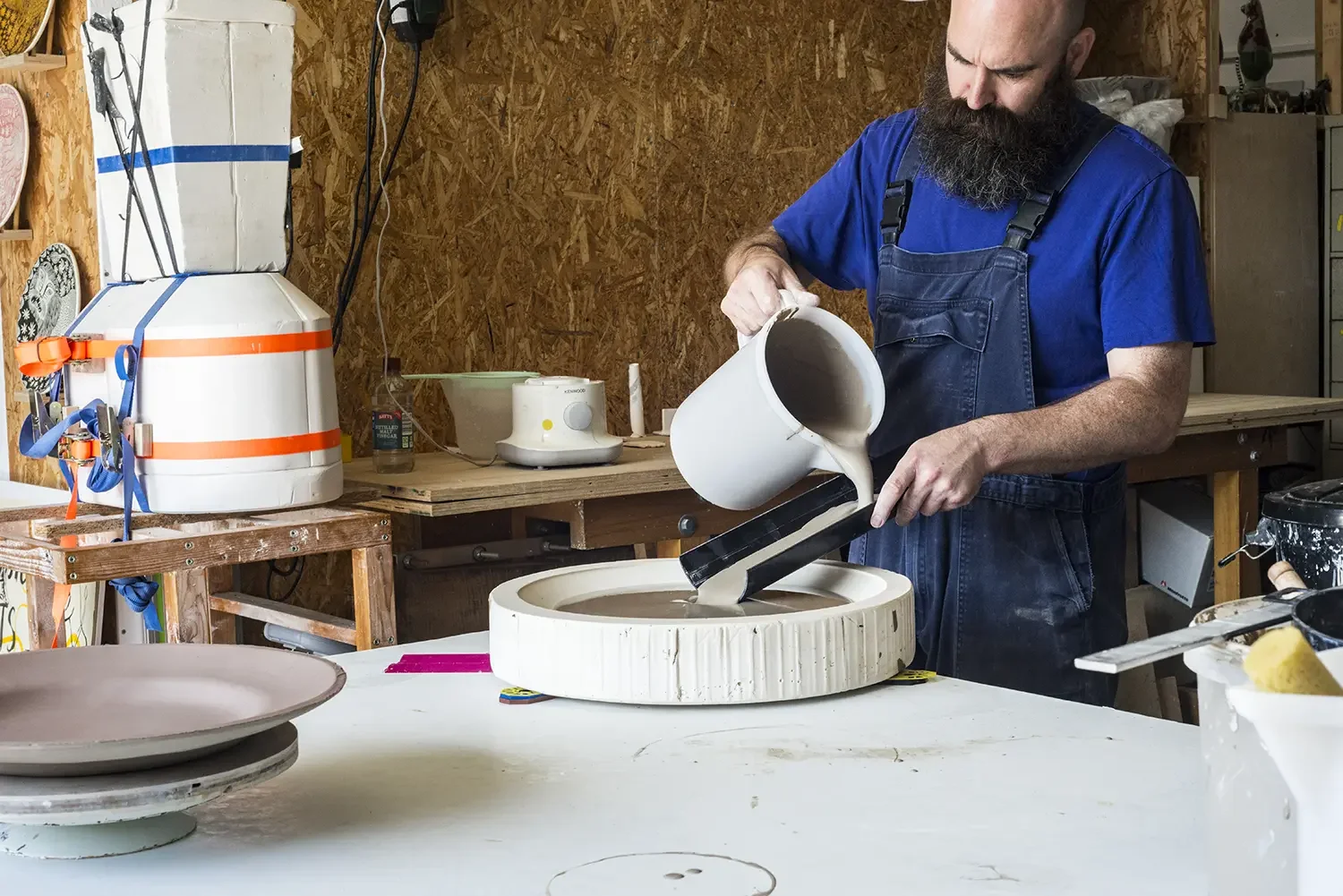Bill Brookes who has a beard and is wearing a blue t-shirt and overalls. He is pouring liquid clay into a circular mould in a pottery workshop.