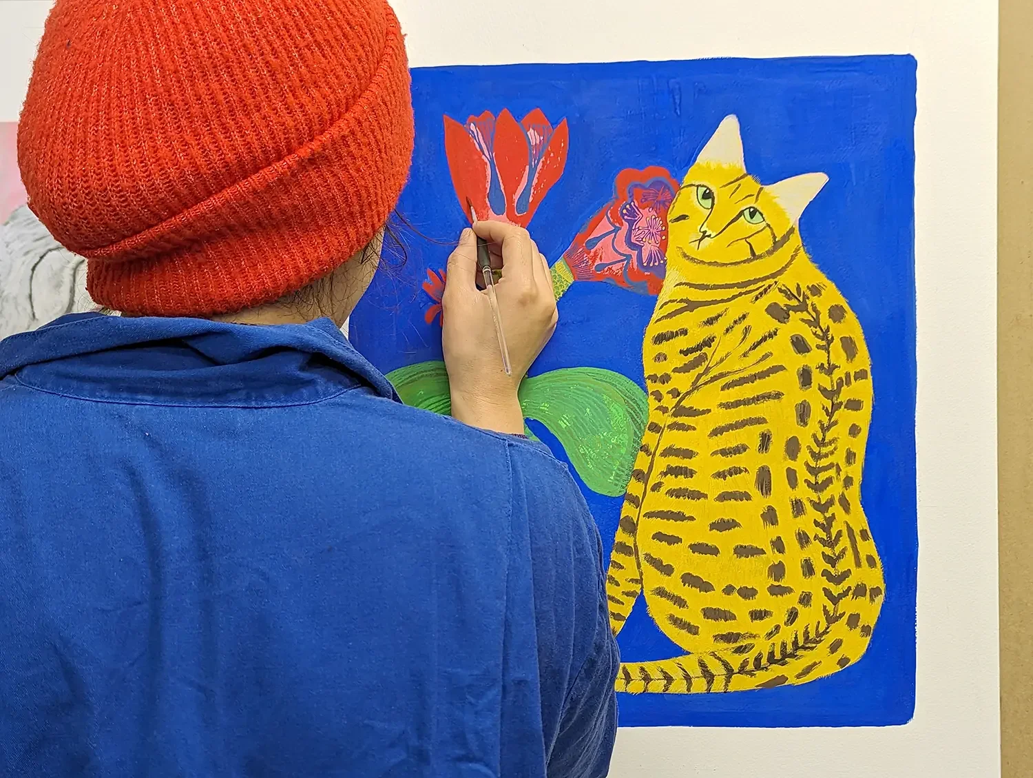 Vicky Lindo wearing a red knit hat and a blue overalls is painting a colourful picture of a yellow cat with black stripes, a pink and purple flower, and green leaves on a blue background.