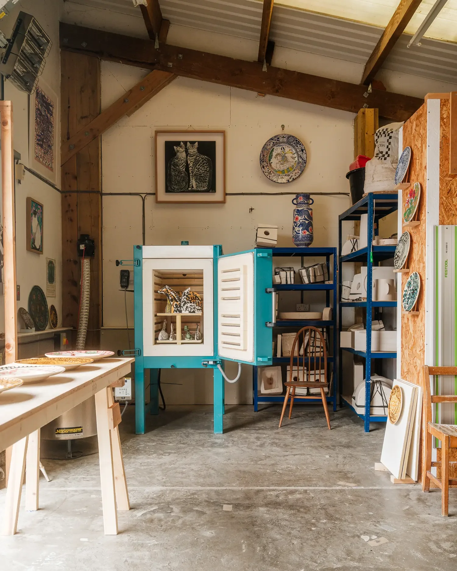 Vicky Lindo and Bill Brooke's art studio with shelves of ceramics, framed artwork, and pottery displayed, with a wooden work table in the foreground and kiln in the background.